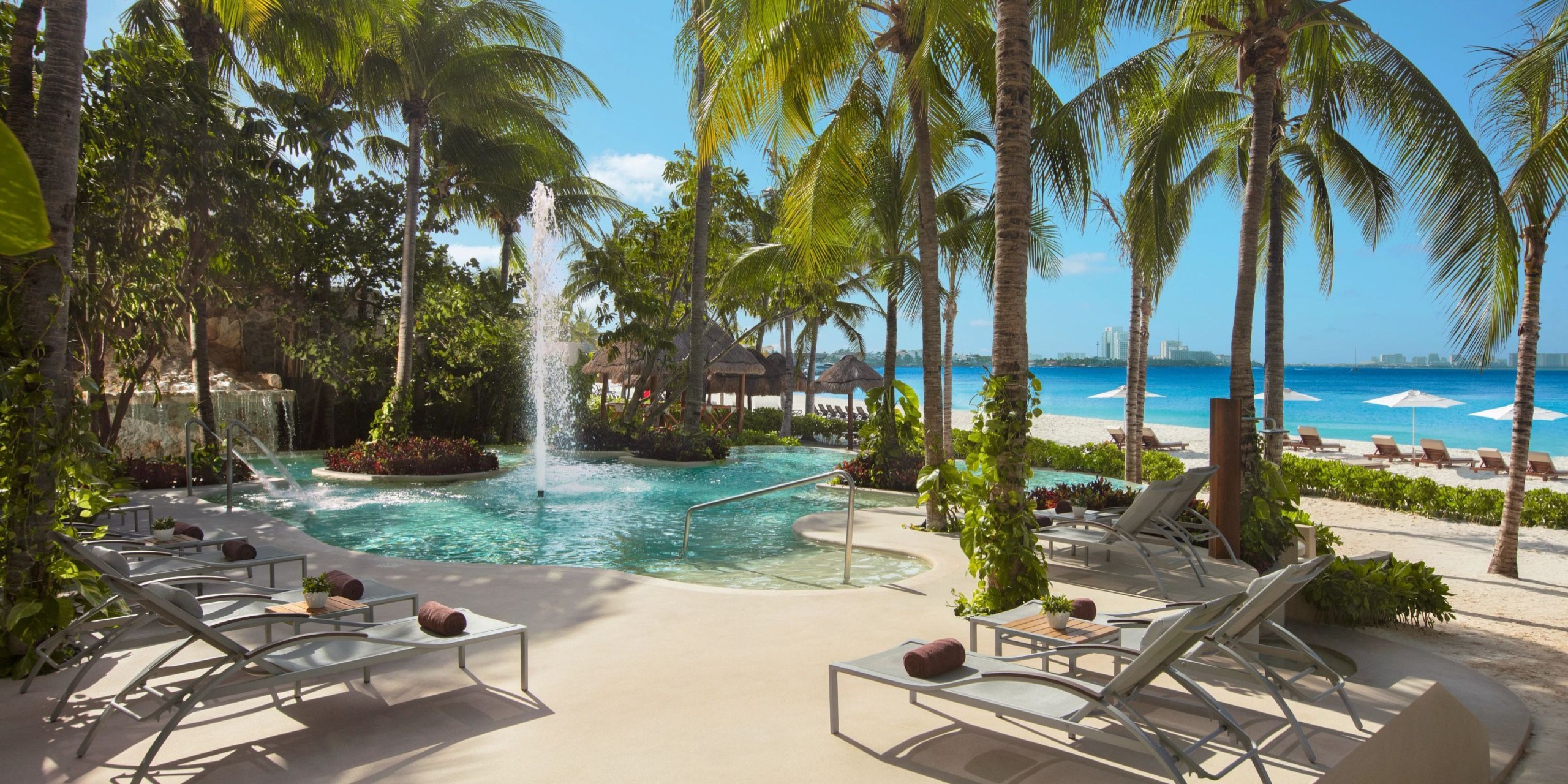 Experience the Dreams Sands Cancun resort, featuring a stunning poolside area adorned with palm trees, sun loungers, and a charming water fountain, all set against the backdrop of a sandy beach and breathtaking ocean views.