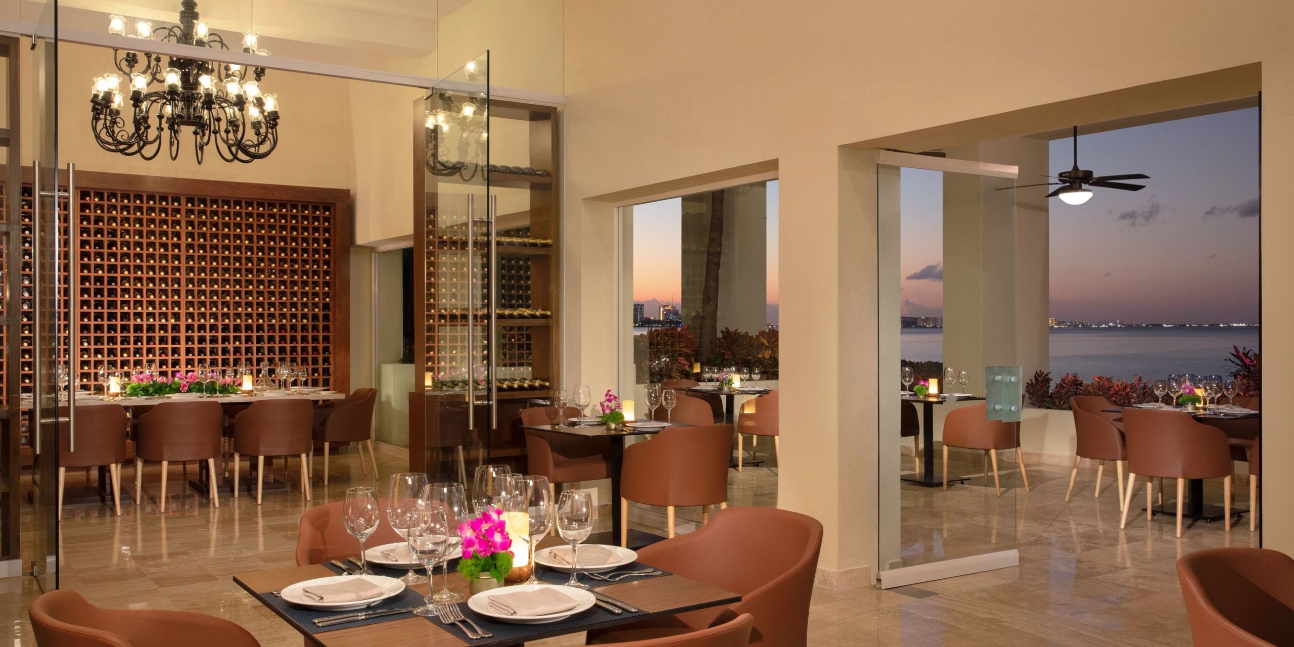 At the Elegant Dreams Sands Cancun restaurant, a stunning chandelier illuminates the area, complemented by a wine rack and tables adorned with glasses and pink flowers. Expansive windows offer a breathtaking view of the sunset over the water.
