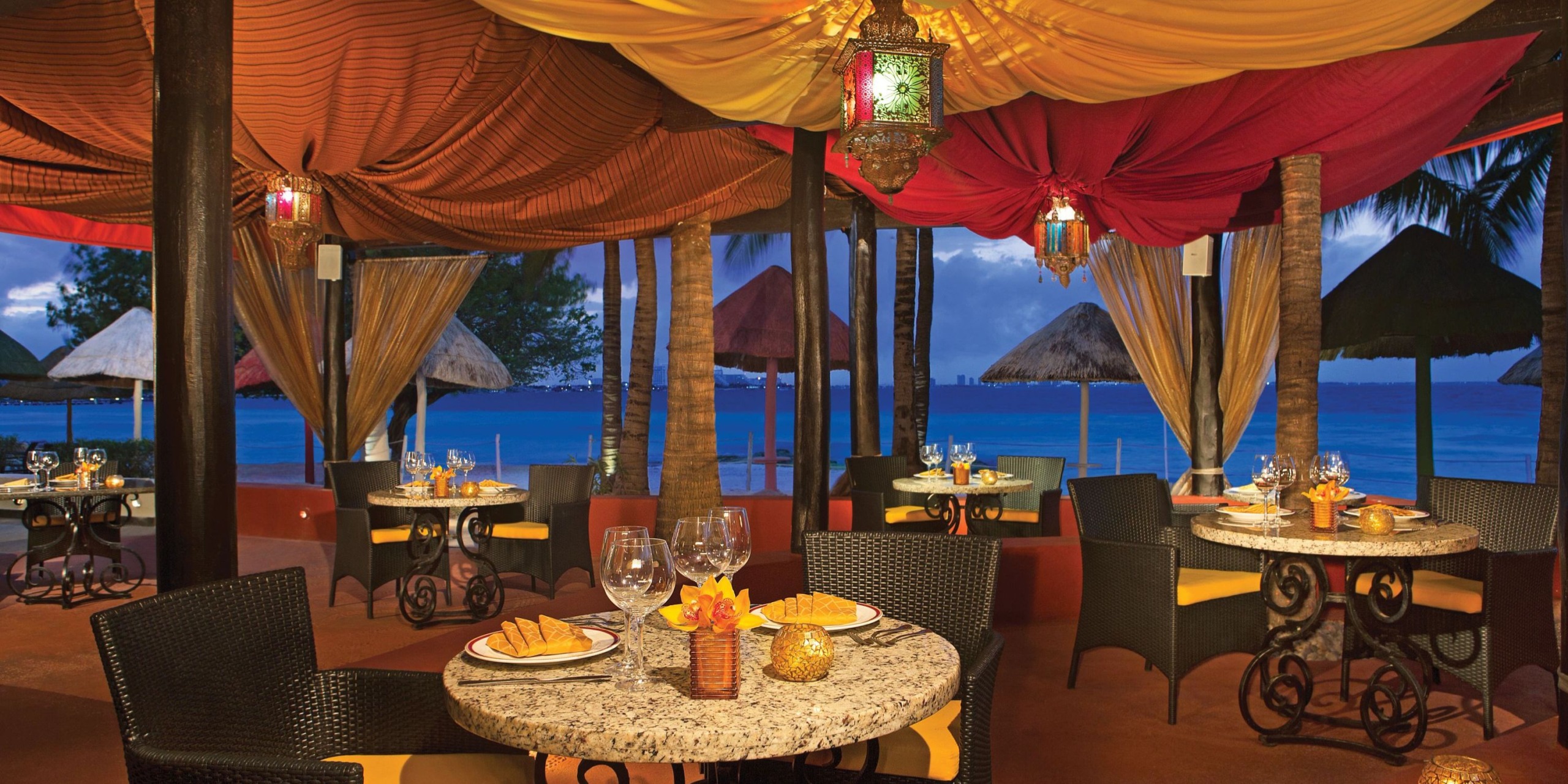 Dreams Sands Cancun features an outdoor dining area overlooking the sea, adorned with colorful drapes and lanterns. The tables are elegantly set with plates, wine glasses, and candles, while palm trees and thatched umbrellas enhance the scenic view.