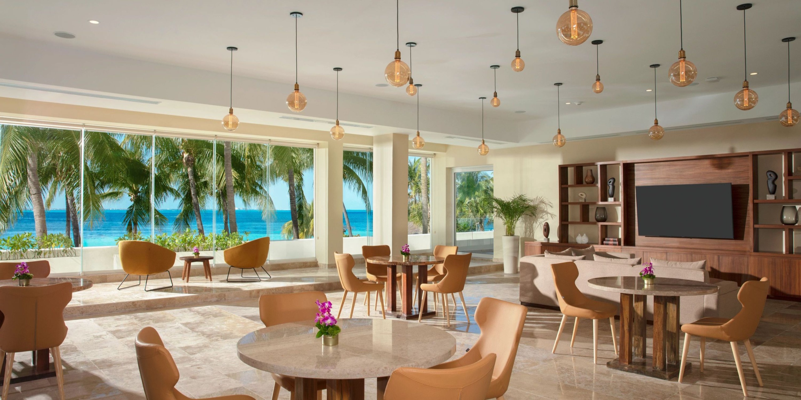 Experience a contemporary, open lounge area at Dreams Sands Cancun, featuring tables and chairs, large windows that provide stunning views of palm trees and the ocean, all illuminated by elegant pendant lights hanging from the ceiling.