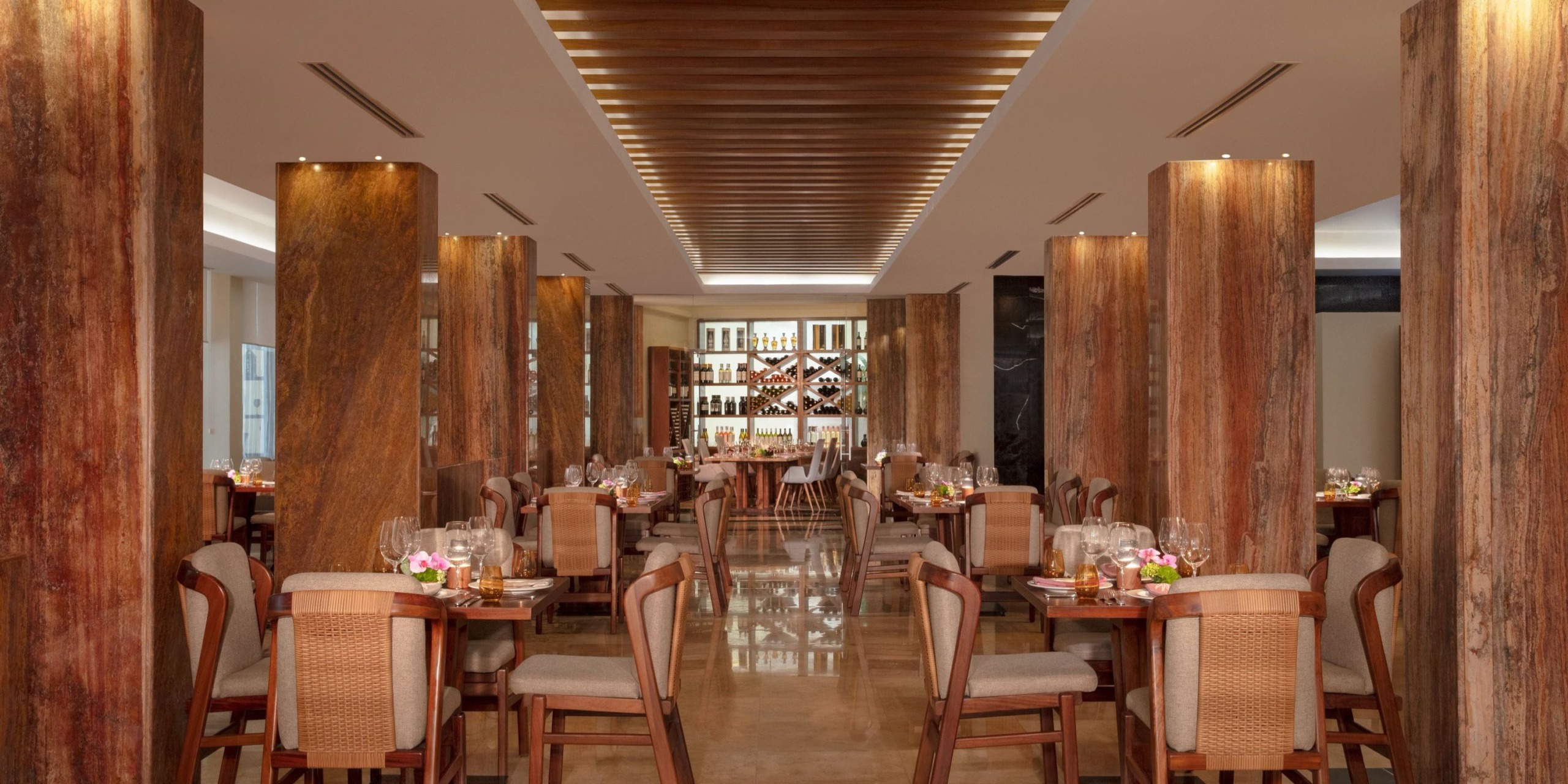 The Dreams Sands Cancun features a dining area with wooden columns, symmetrically arranged tables and chairs, and at the far end, there's a wine rack along with shelves.