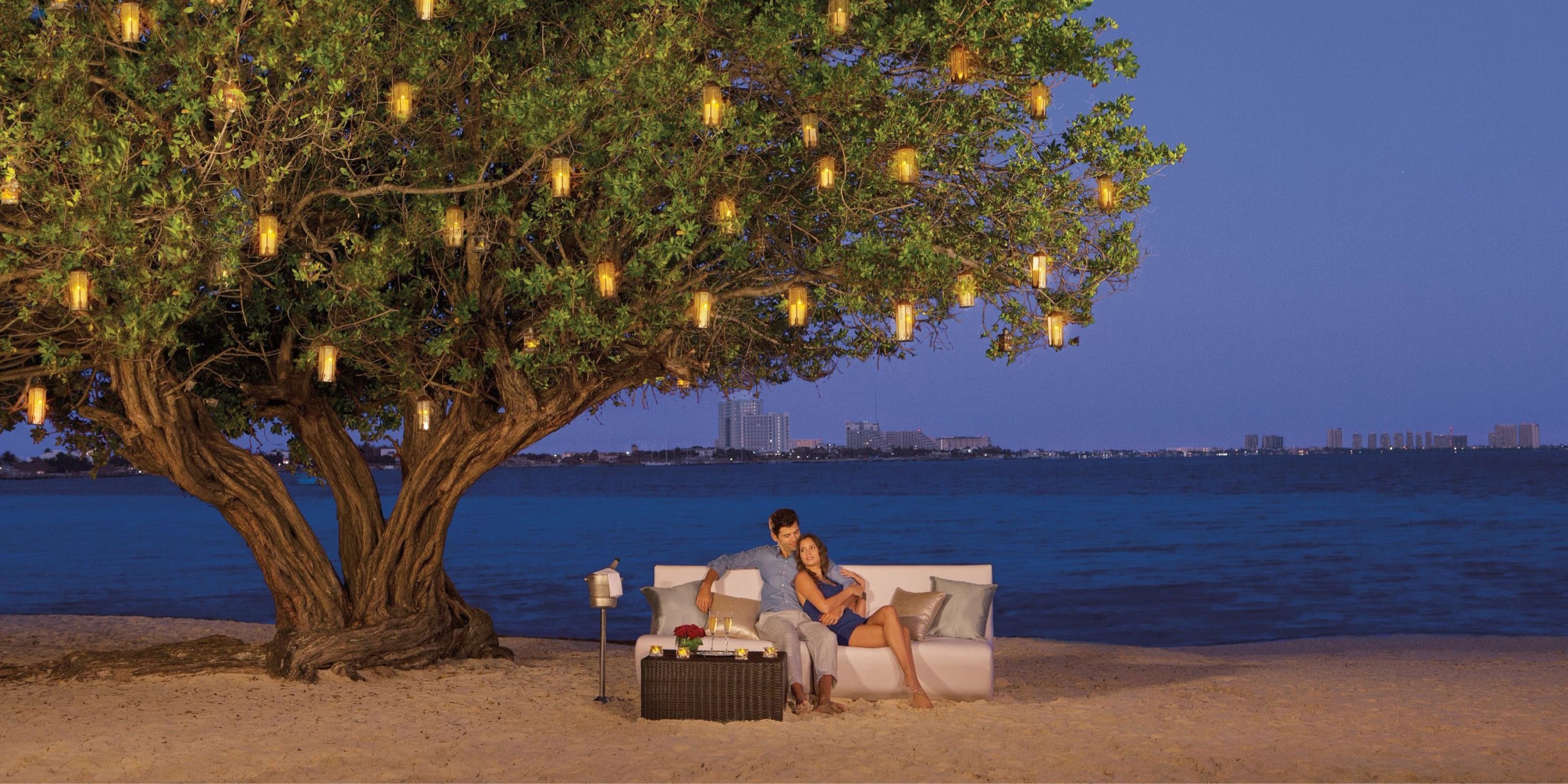 A couple relaxes on the beach at Dreams Sands Cancun, seated beneath a large tree adorned with twinkling lights next to a table where a lit candle and drinks rest. The city skyline beautifully frames the water as dusk approaches.
