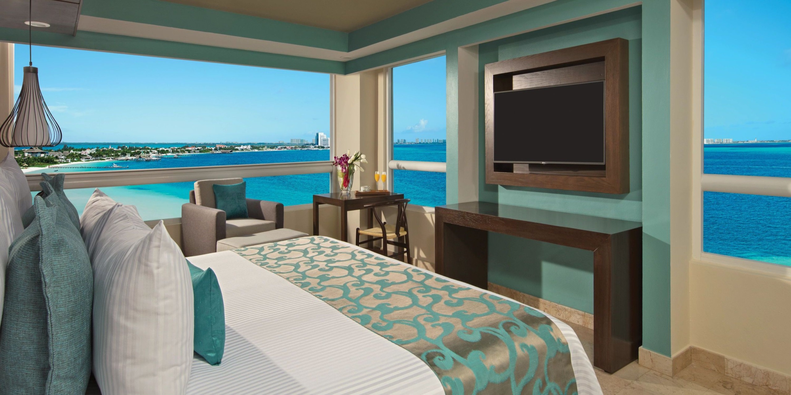 Hotel room at Dreams Sands Cancun featuring large windows with stunning ocean and city views. Includes a bed adorned with teal and white bedding, a cozy armchair, a small table, and a wall-mounted TV.