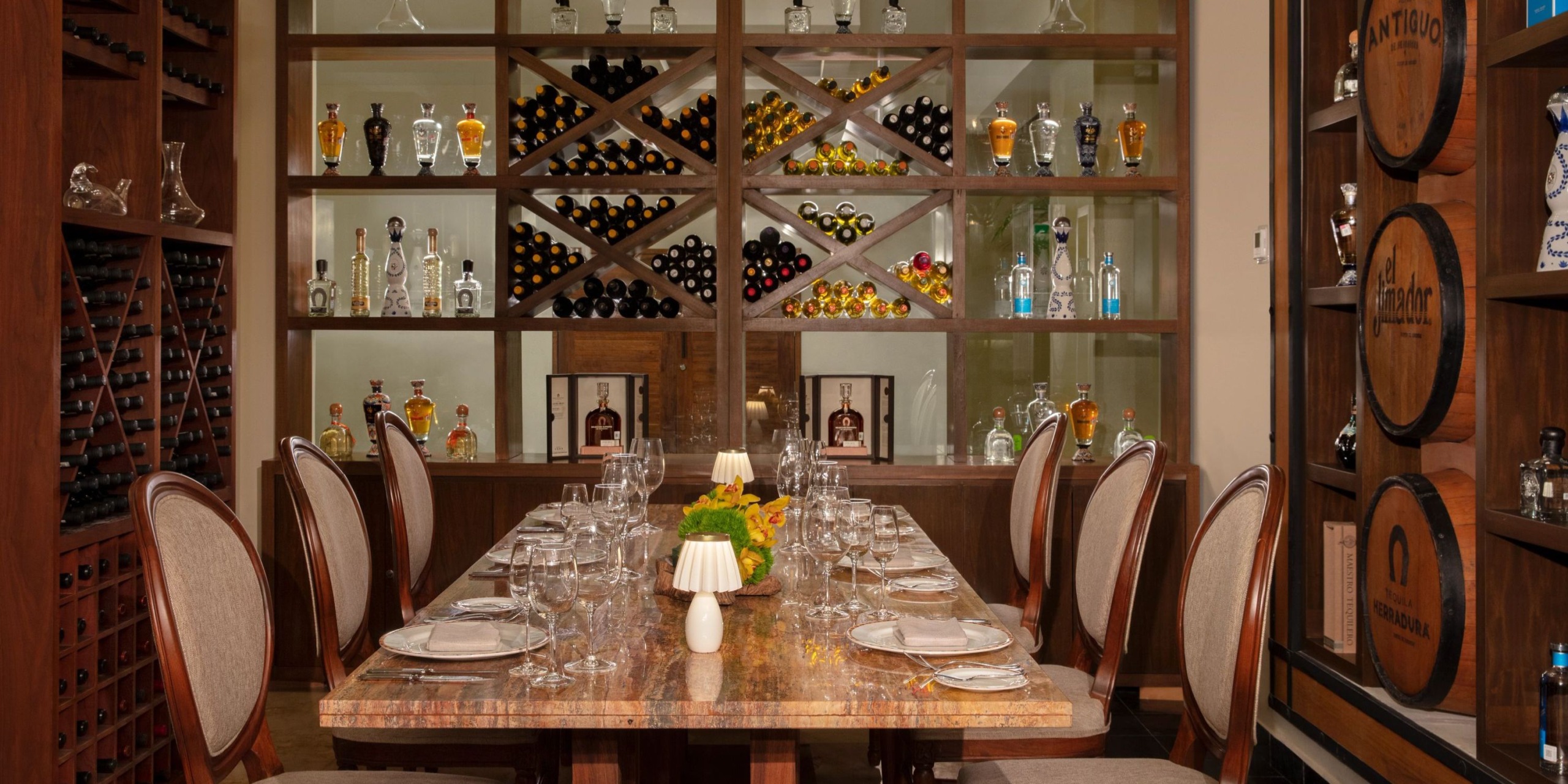 The luxurious dining room at Dreams Sands Cancun features a beautifully set long table, surrounded by stylish wooden chairs. Shelves in the background are adorned with an assortment of wine bottles and gleaming glassware.