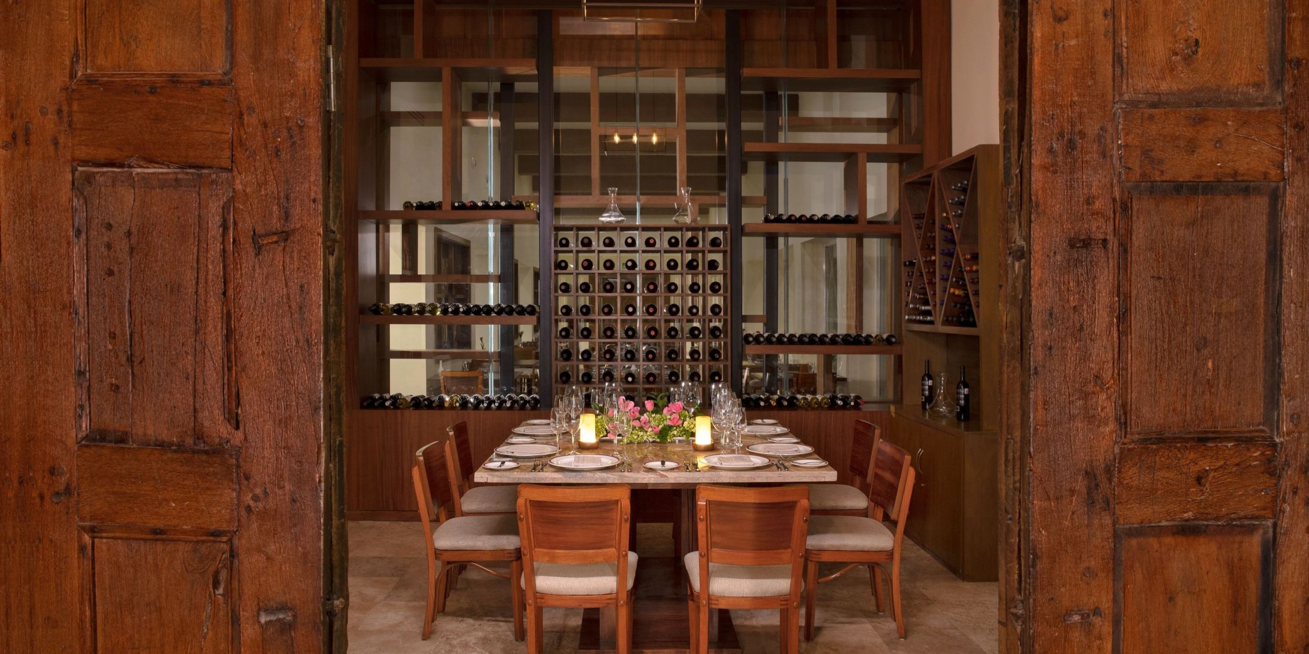 A dining room at Dreams Sands Cancun, featuring a wooden table set for eight and surrounded by shelves of wine bottles. The table is beautifully decorated with candles and a floral centerpiece, while wooden doors frame the view.