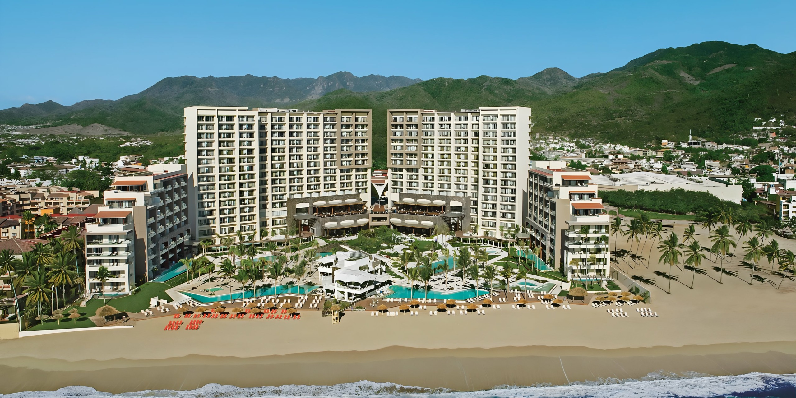 Dreams Vallarta Bay beachfront resort with swimming pools flanked by a wedding venue in a twin-building structure and surrounded by mountains.