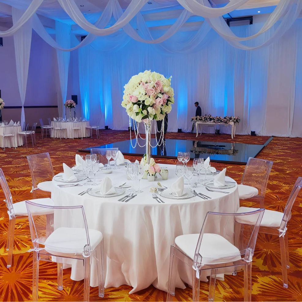 A round table set for an event with white tablecloth, floral centerpiece, and glassware in a large hall with draped ceiling and blue uplighting. Clear chairs surround the table.
