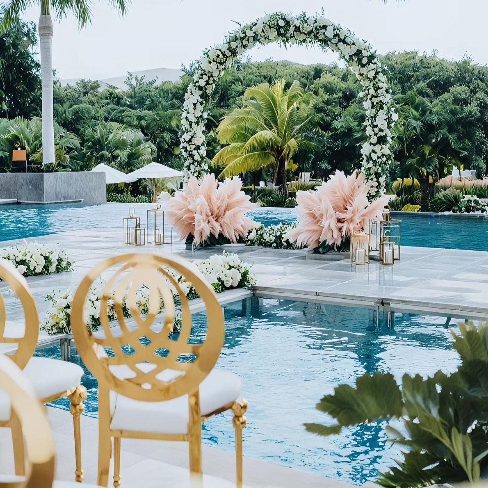 An elegant wedding setup by a pool with golden chairs, a floral arch, and pink feathery decorations surrounded by lush greenery.