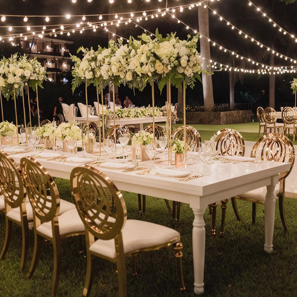 An outdoor wedding reception setup featuring elegantly decorated white tables, high floral arrangements, gold chairs, and string lights illuminating the scene.