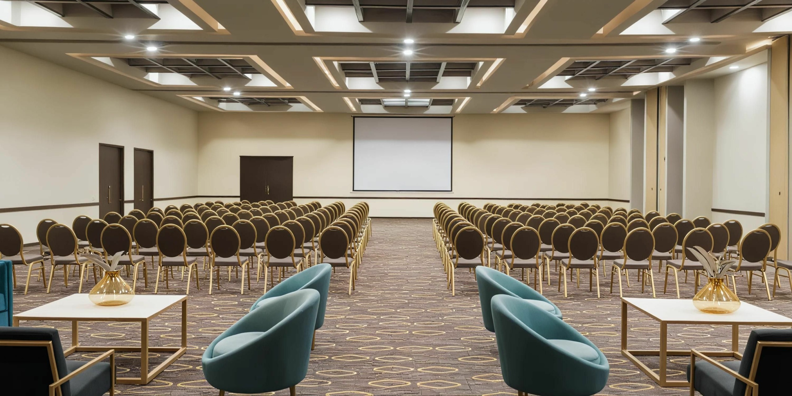 A conference room with rows of empty chairs facing a large projection screen. Four teal chairs and white tables are positioned at the front of the room.