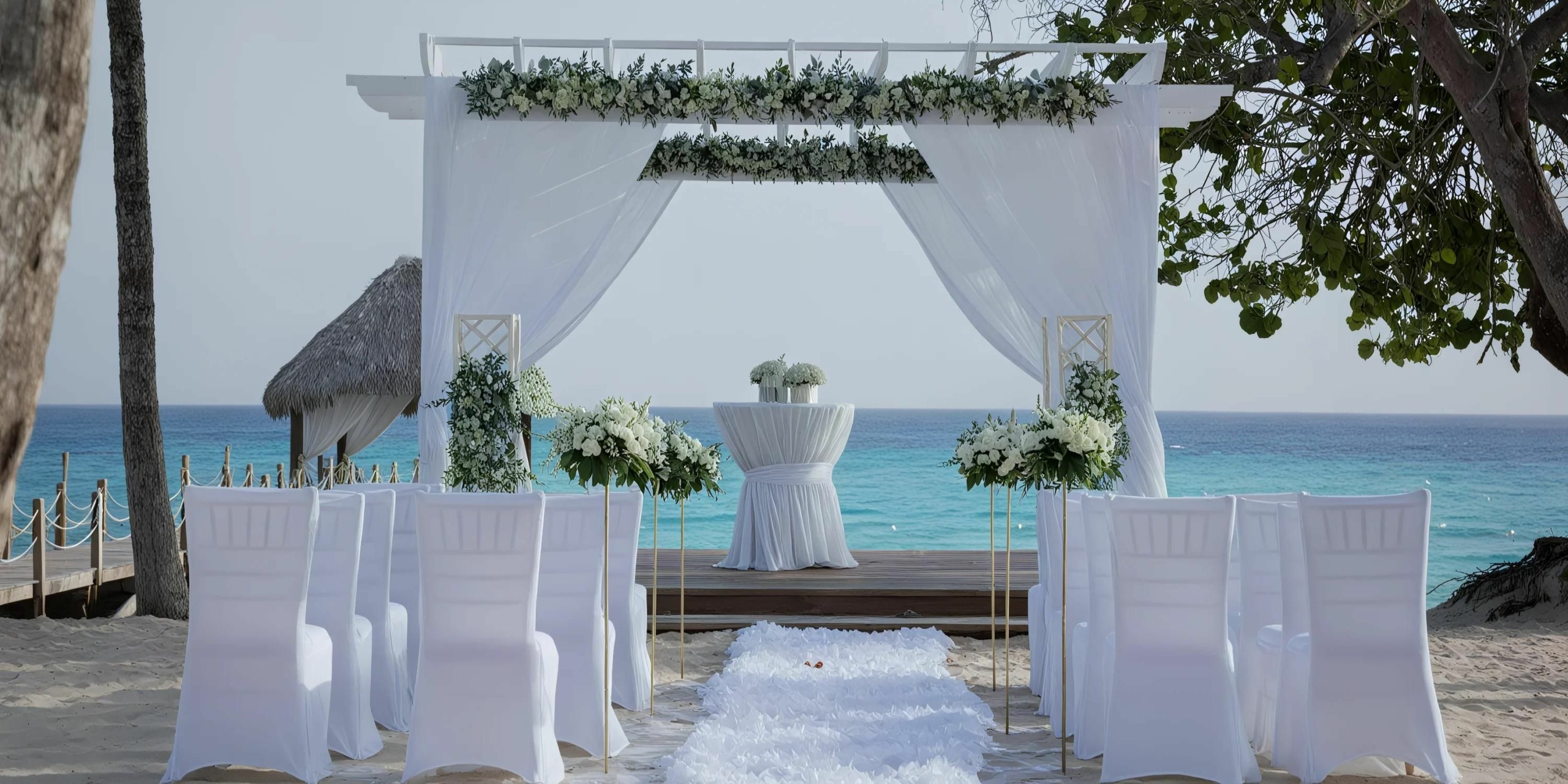 A white outdoor wedding altar is set up on a sandy beach, overlooking the ocean. White chairs and floral arrangements line the aisle leading to the arch, which is decorated with white drapery and flowers.