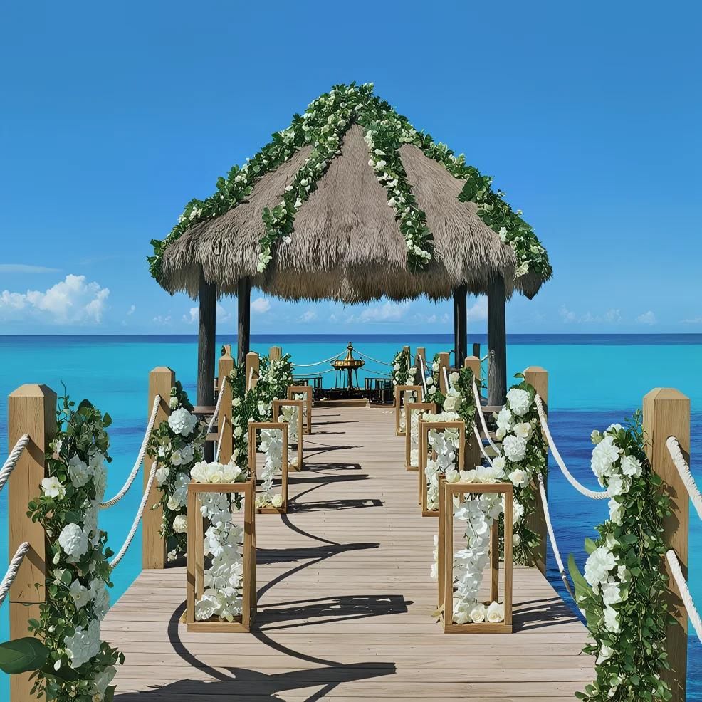 A wooden pier decorated with white flowers and greenery leads to a thatched-roof gazebo over turquoise ocean water under a clear blue sky.