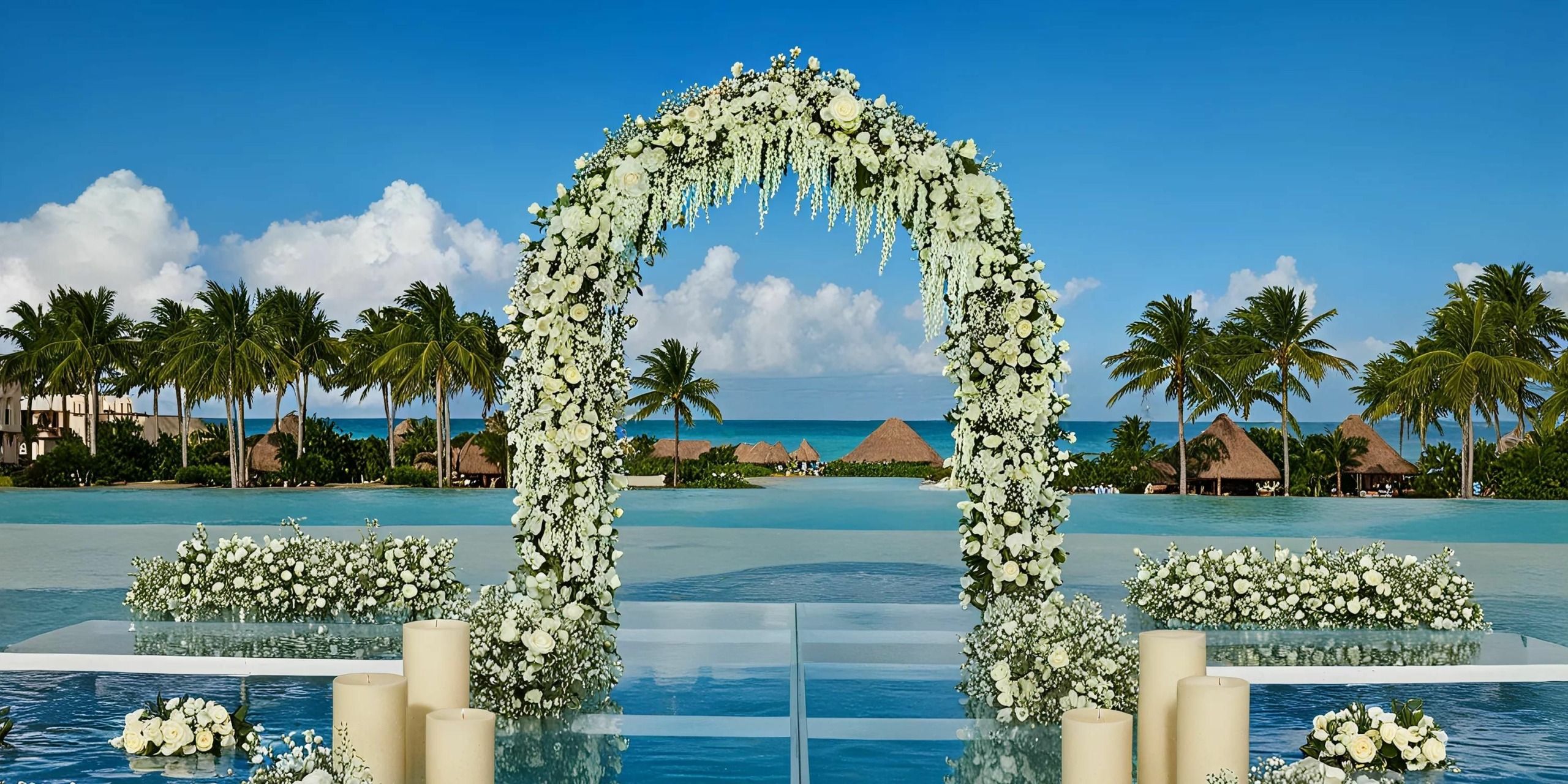 A white floral arch stands on a platform over water, with palm trees, thatched roofs, and the ocean in the background under a blue sky. Cream-colored candles are placed around the arch.