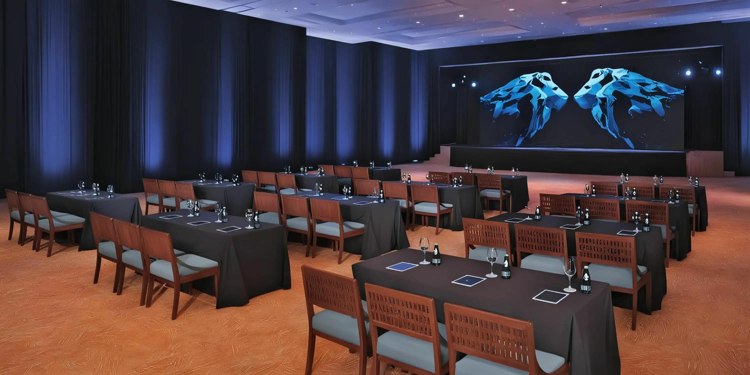 A conference room setup with tables and chairs facing a stage illuminated by blue lights and adorned with an abstract blue and white artwork at Dreams Sands Cancun. The tables are equipped with stationery and water bottles.