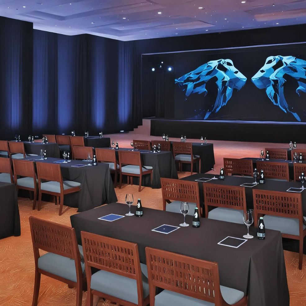 A conference room set up with black-clothed tables, wooden chairs, and place settings for attendees. A large screen displaying abstract blue shapes is at the front of the room.