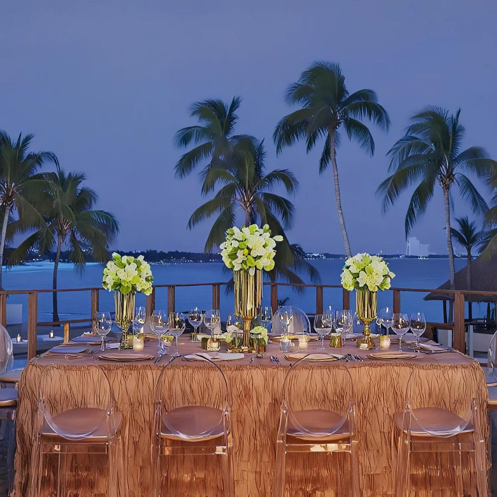 Elegant outdoor dining table set with floral centerpieces and glassware, with a backdrop of palm trees and a calm ocean at dusk.