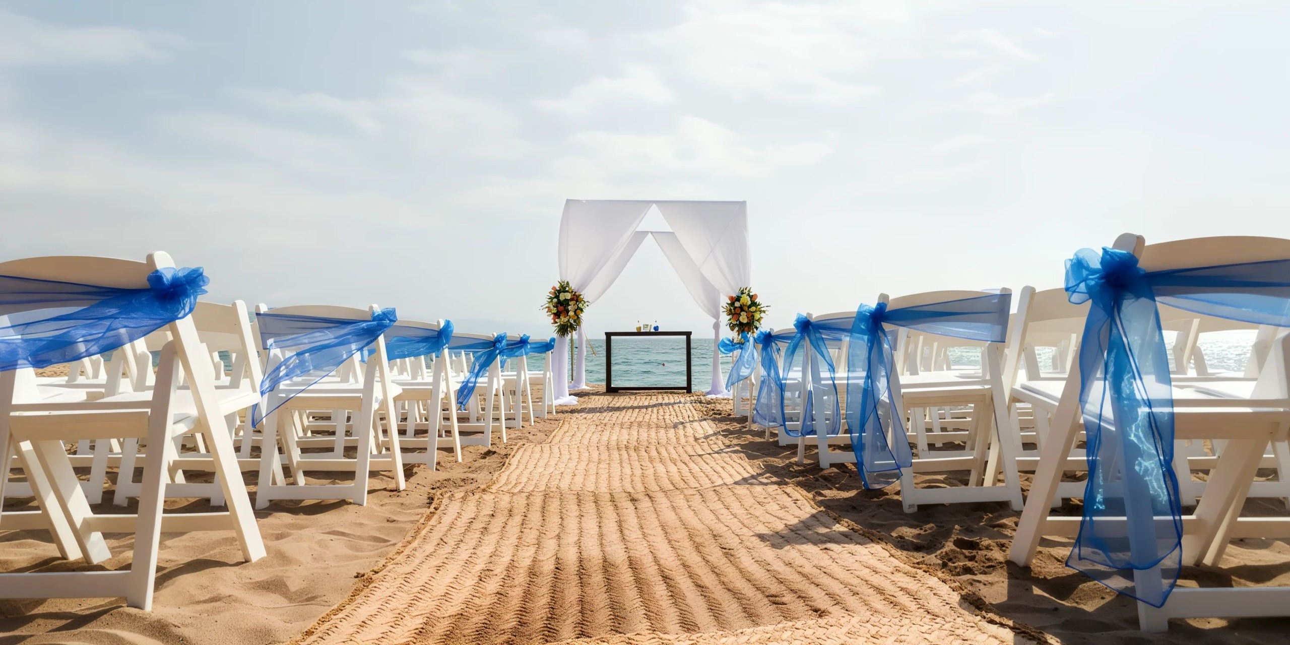 Beach wedding setup with a sandy aisle leading to an altar adorned with white fabric and flowers. White chairs with blue ribbons are arranged on either side.