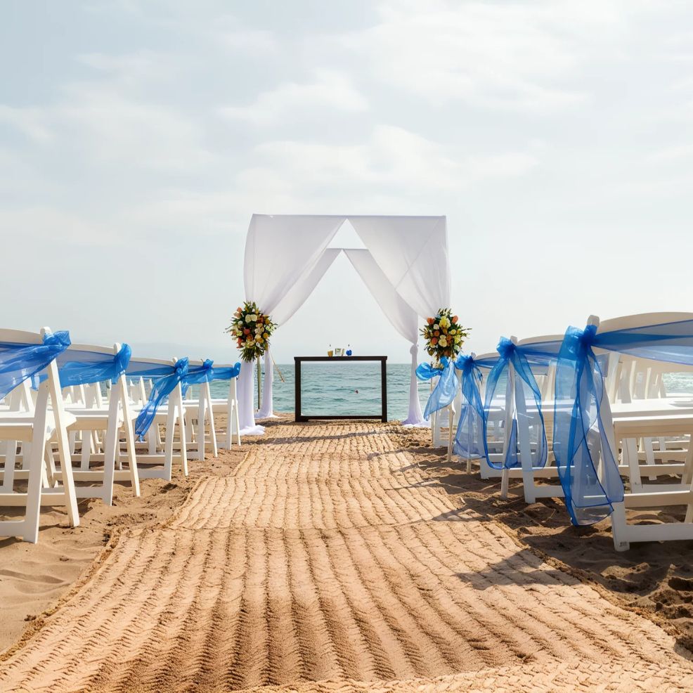 Beach wedding setup with white chairs and blue ribbons, a sandy aisle leading to an altar adorned with flowers, and the ocean in the background.