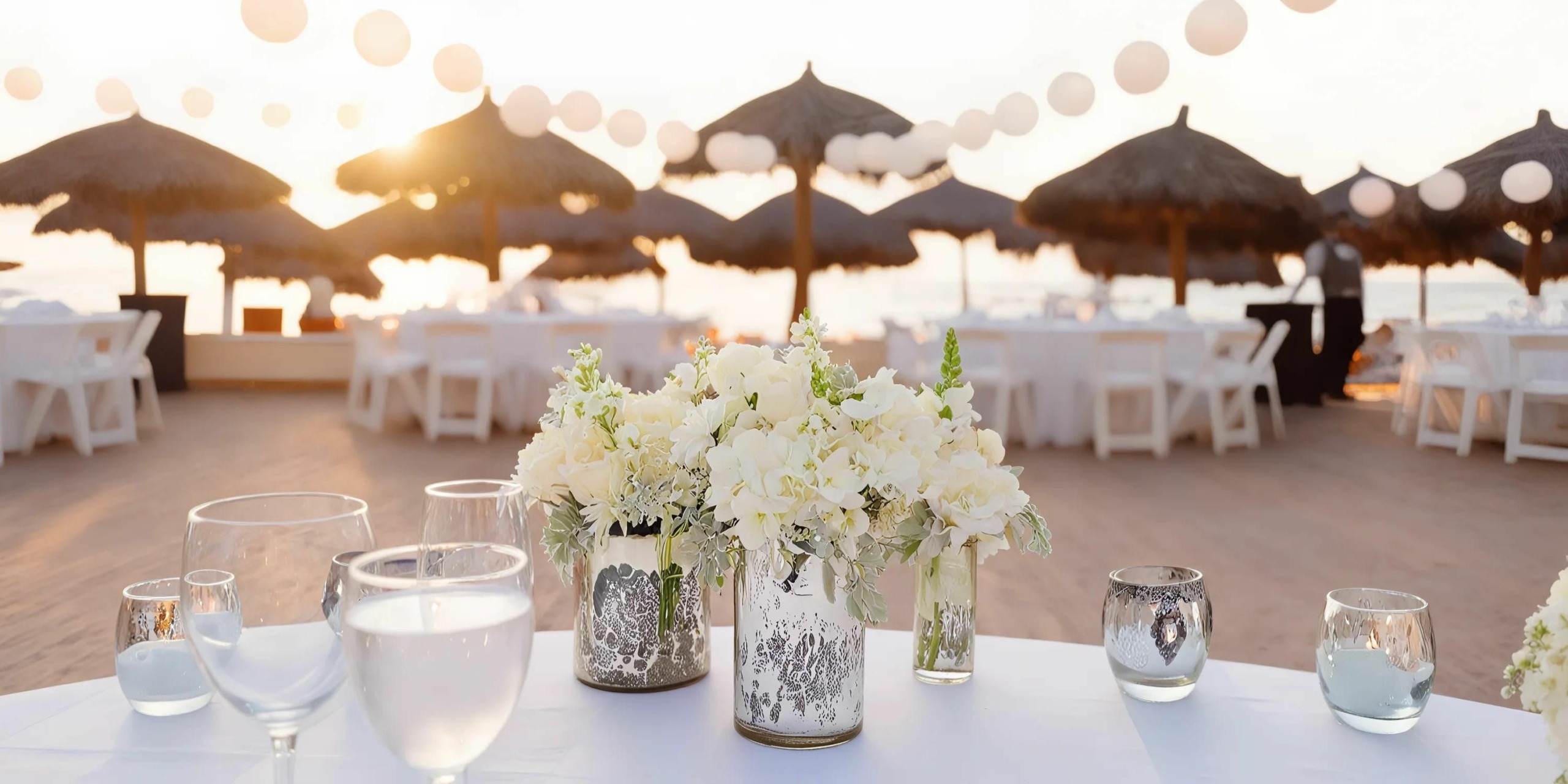 An outdoor event setting with white floral arrangements, glassware on tables, and thatched-roof umbrellas at sunset.