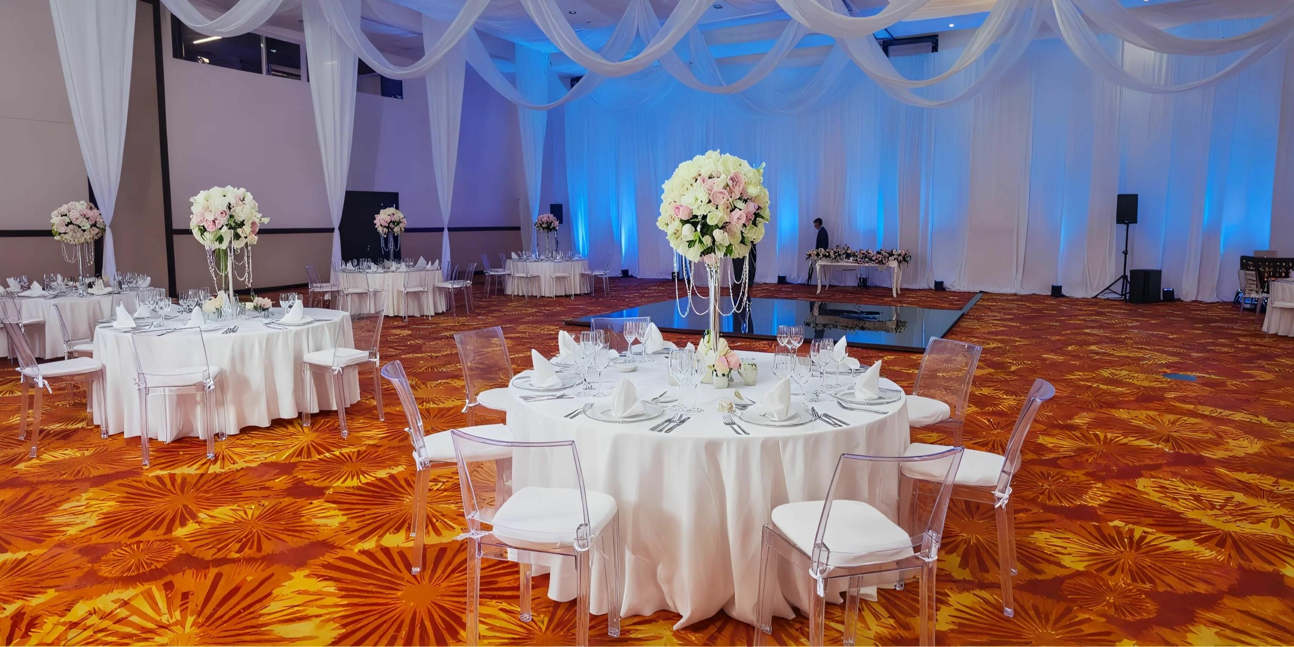 A decorated event hall with round tables covered in white cloths, floral centerpieces, clear chairs, and a stage with blue lighting. Draped fabric hangs from the ceiling.
