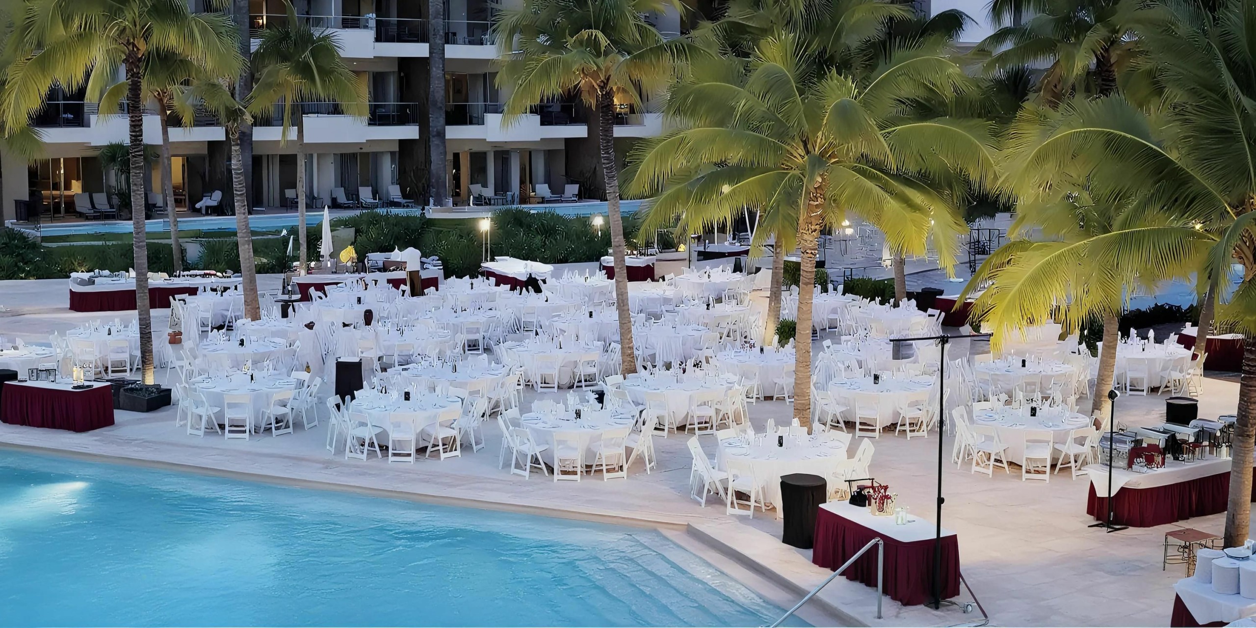 The Dreams Vallarta Bay poolside area is set up with numerous round tables covered in white tablecloths and surrounded by white chairs for an outdoor event, with palm trees and hotel balconies visible in the background.