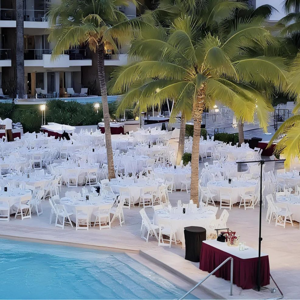 Outdoor dining area by a pool with numerous white-clothed tables and chairs surrounded by palm trees.
