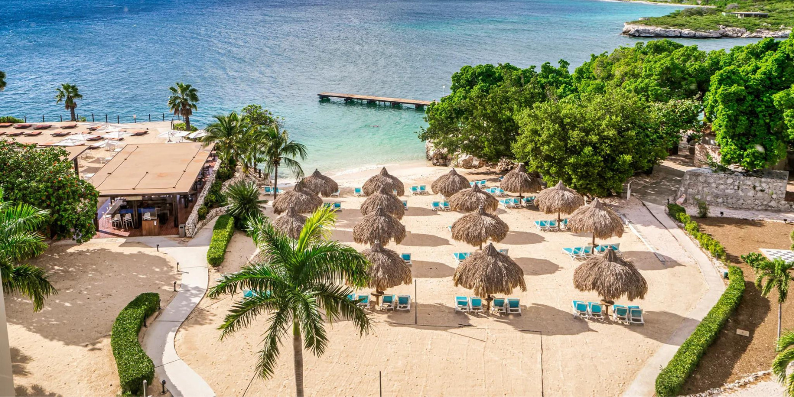 A seaside resort with thatched-roof umbrellas, sun loungers, a sandy beach, clear turquoise waters, lush greenery, and a dock extending into the sea, Dreams Curacao is perfect for LGBTQ+ friendly destination wedding resorts in the Caribbean Islands.