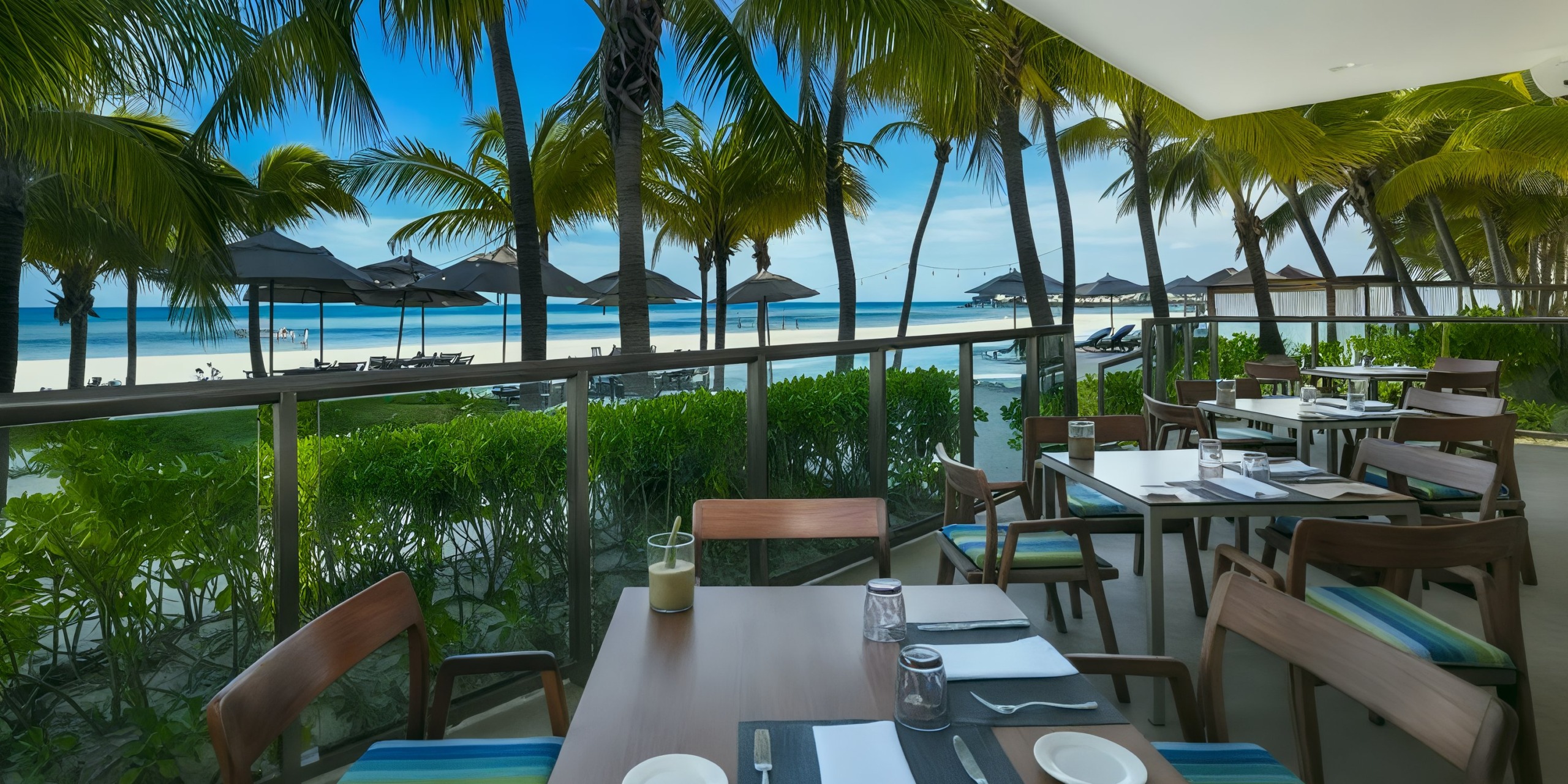 Outdoor dining experience at El Dorado Maroma, featuring wooden tables and striped chairs set beneath palm trees with a stunning beach and ocean view.