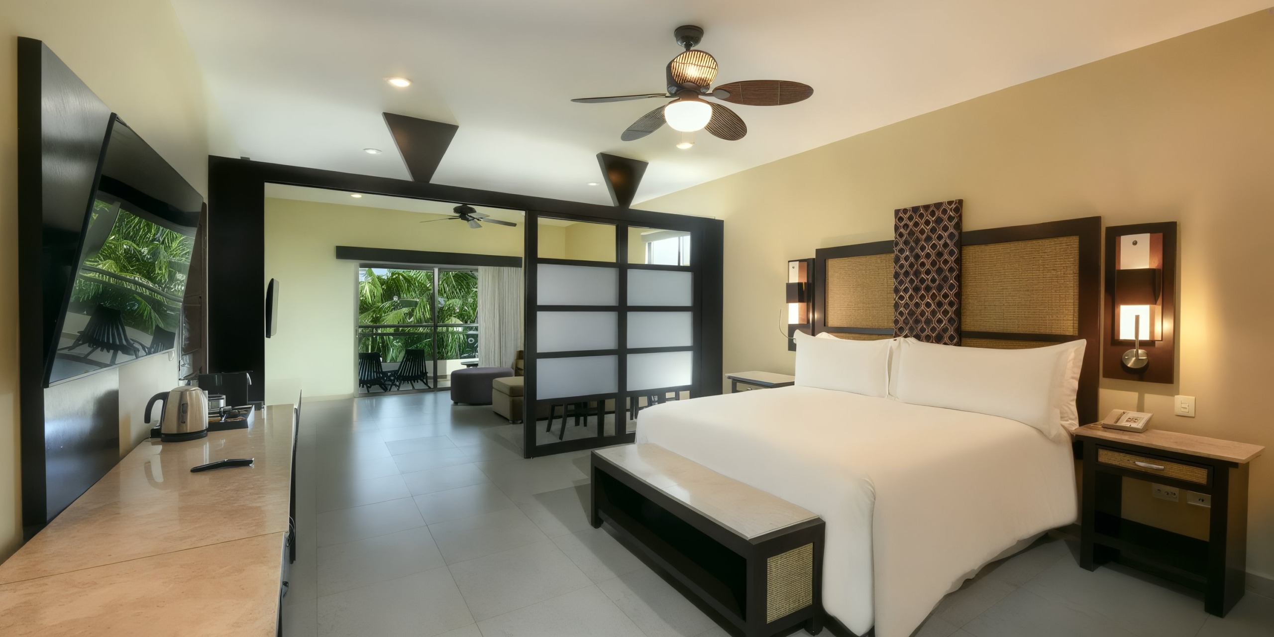 Experience the spacious accommodations at El Dorado Maroma, featuring a king-sized bed, ceiling fan, TV, and a comfortable seating area. Enjoy breathtaking tropical views through large windows.
