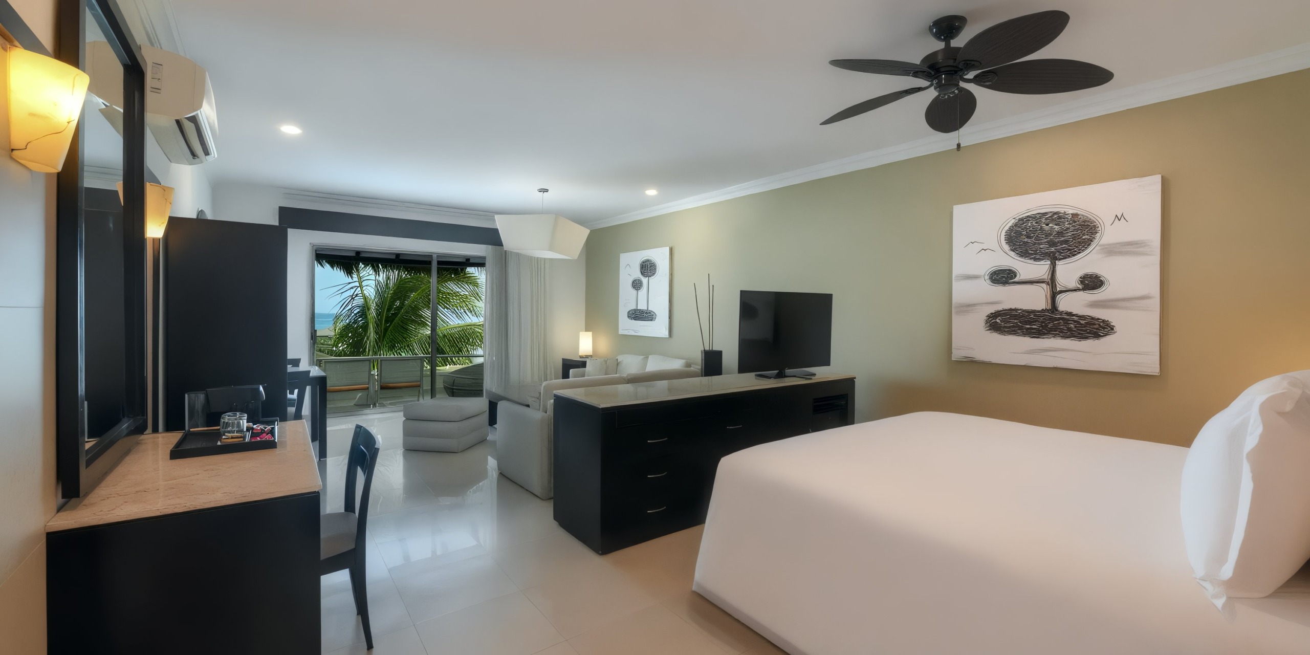 Experience luxury at El Dorado Maroma with a spacious room featuring a large bed, wall-mounted TV, beautiful artwork, and a convenient desk. Enjoy breathtaking views of palm trees and the ocean through the sliding door that leads to your private balcony. The room is elegantly enhanced with a ceiling fan and modern lighting.