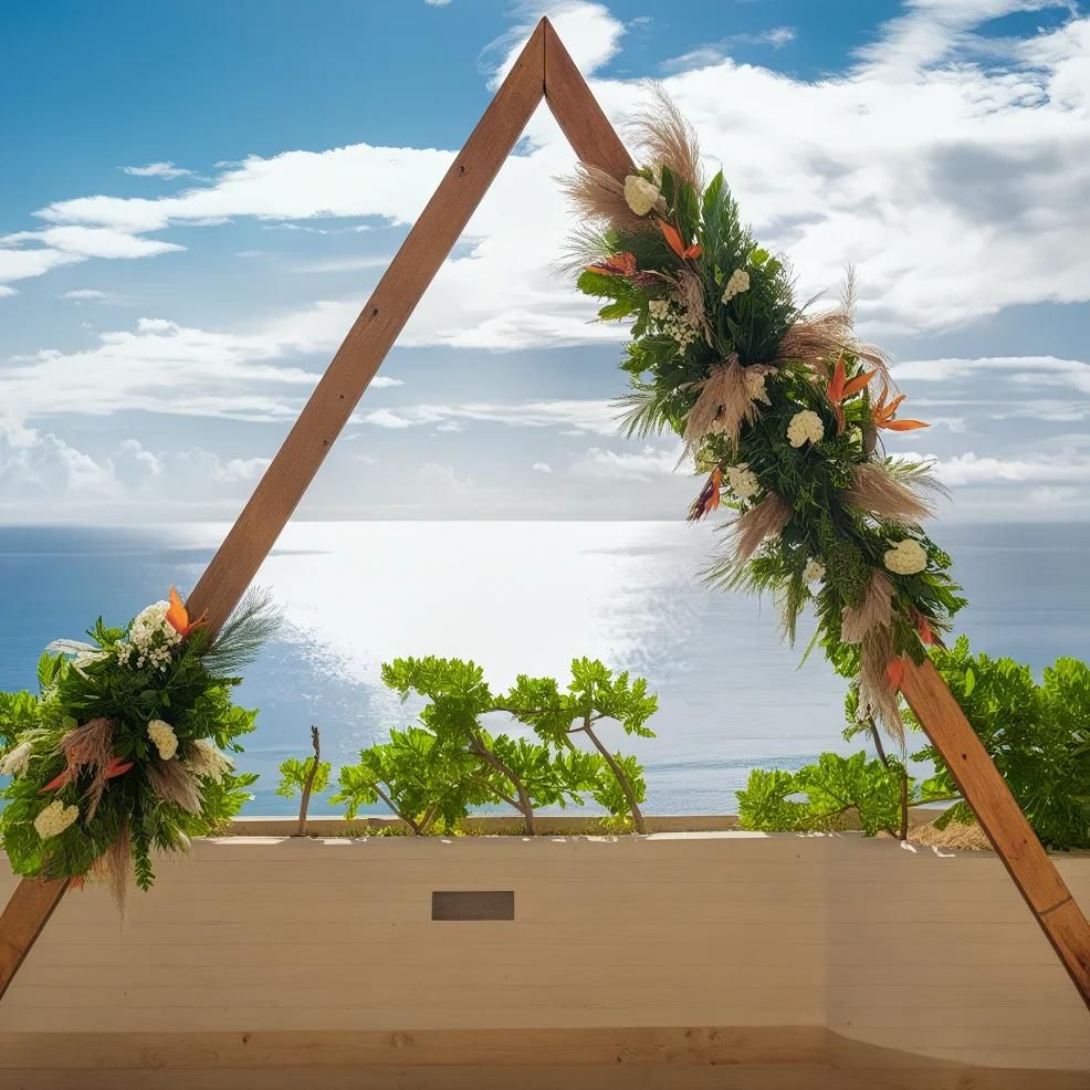 A triangular wooden arch decorated with flowers and greenery stands on a terrace overlooking the ocean under a partly cloudy sky.