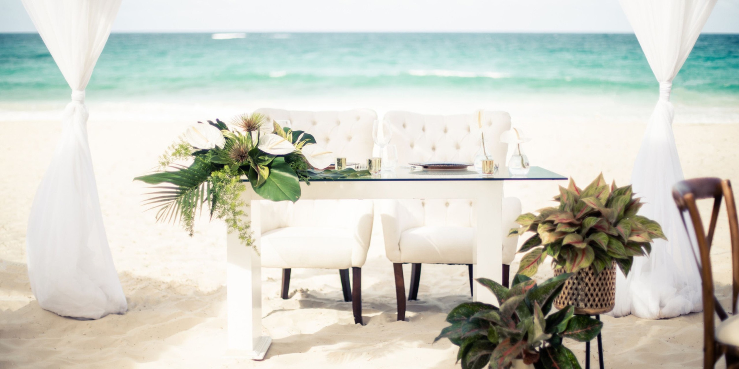 A table with white chairs is set up on the sandy beach at Hard Rock Cancun, surrounded by lush green plants and floral decorations, with the ocean visible in the background.