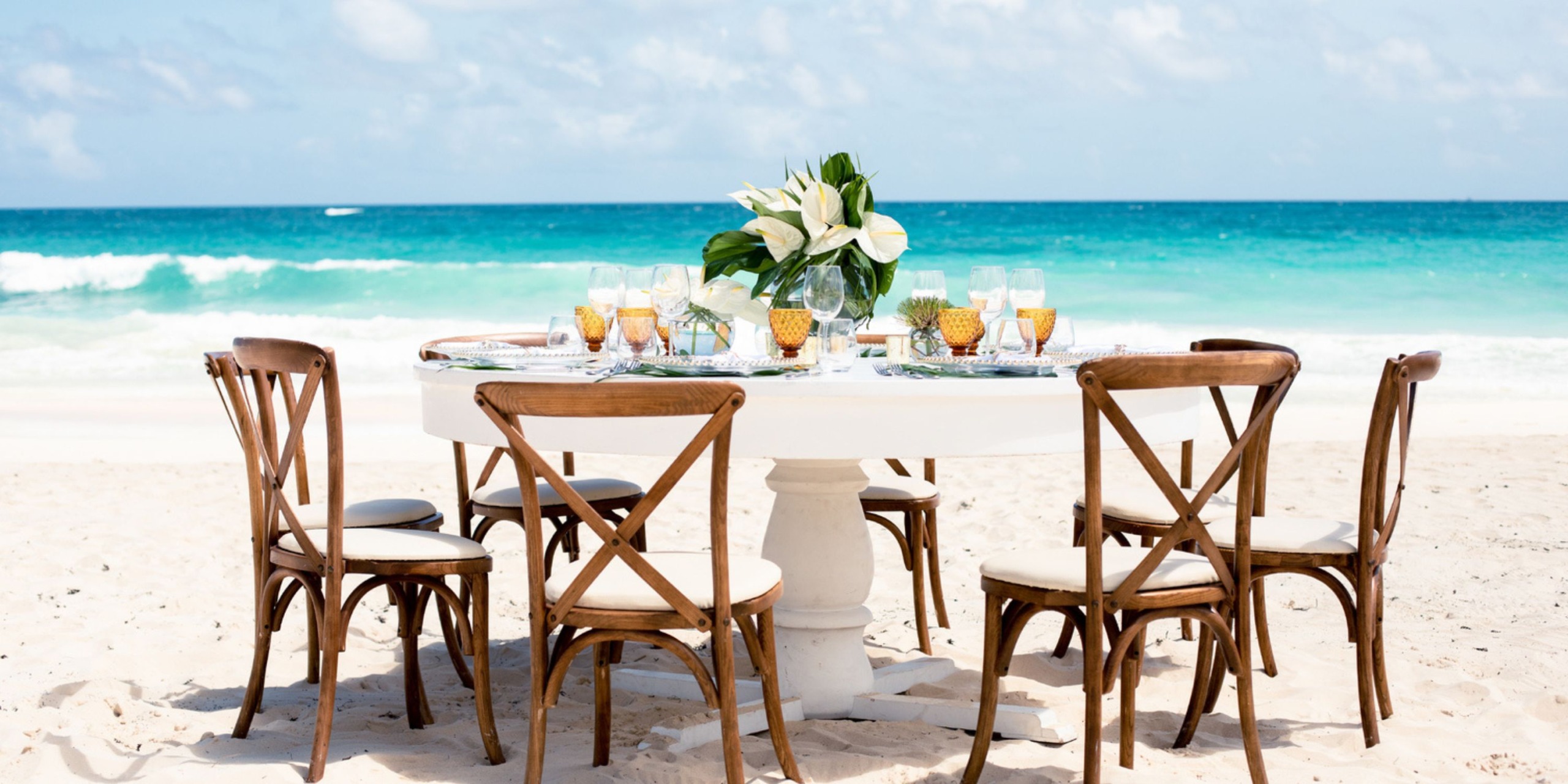 At Hard Rock Cancun, a round table with six wooden chairs graces the sandy beach. It is adorned with plates, glasses, and a vase of white flowers, against a backdrop of ocean and blue sky.