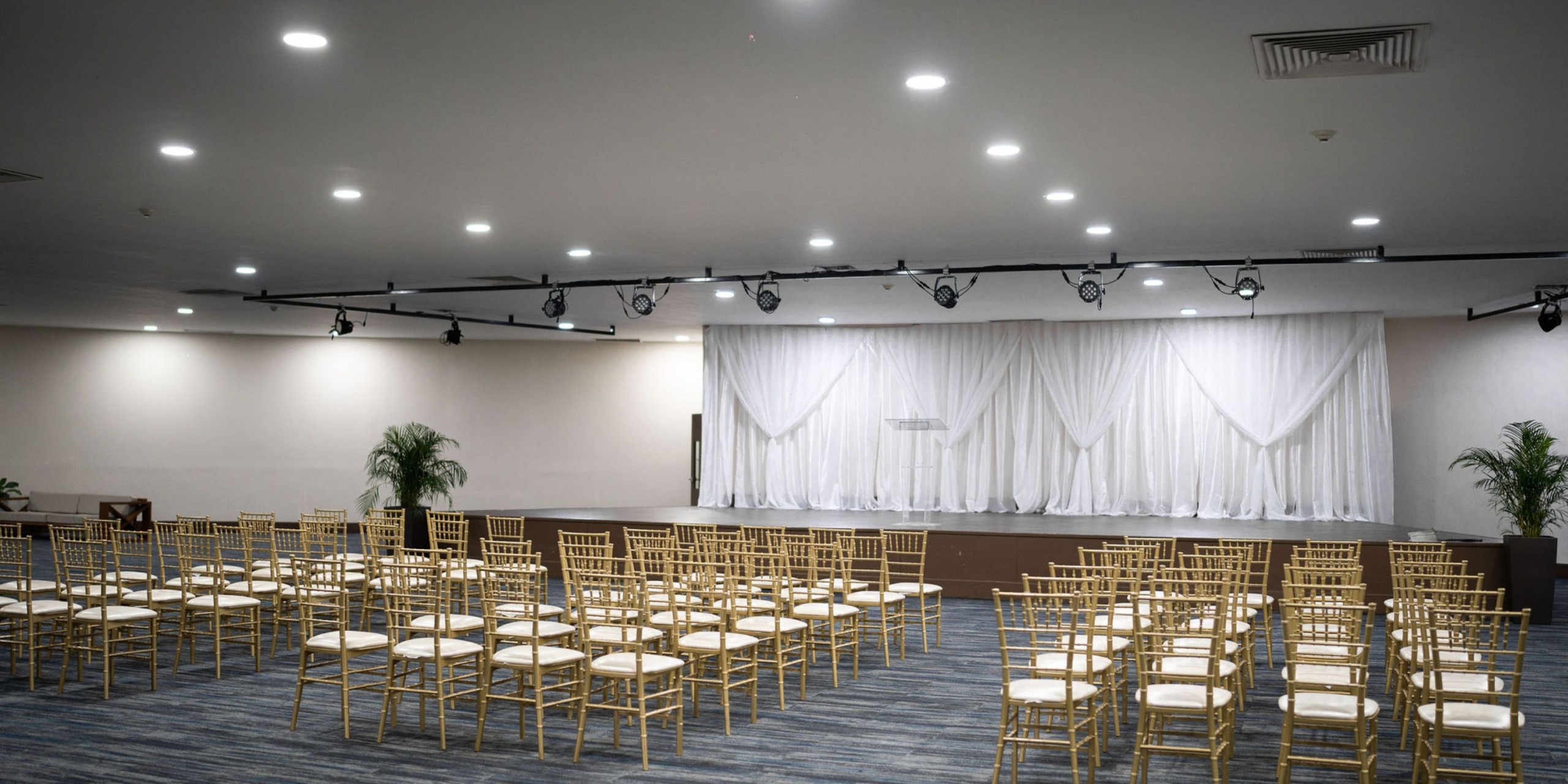 The Hideaway at Royalton Blue Waters offers an event room featuring rows of gold chairs facing a stage adorned with elegant white drapery and illuminated by spotlights.