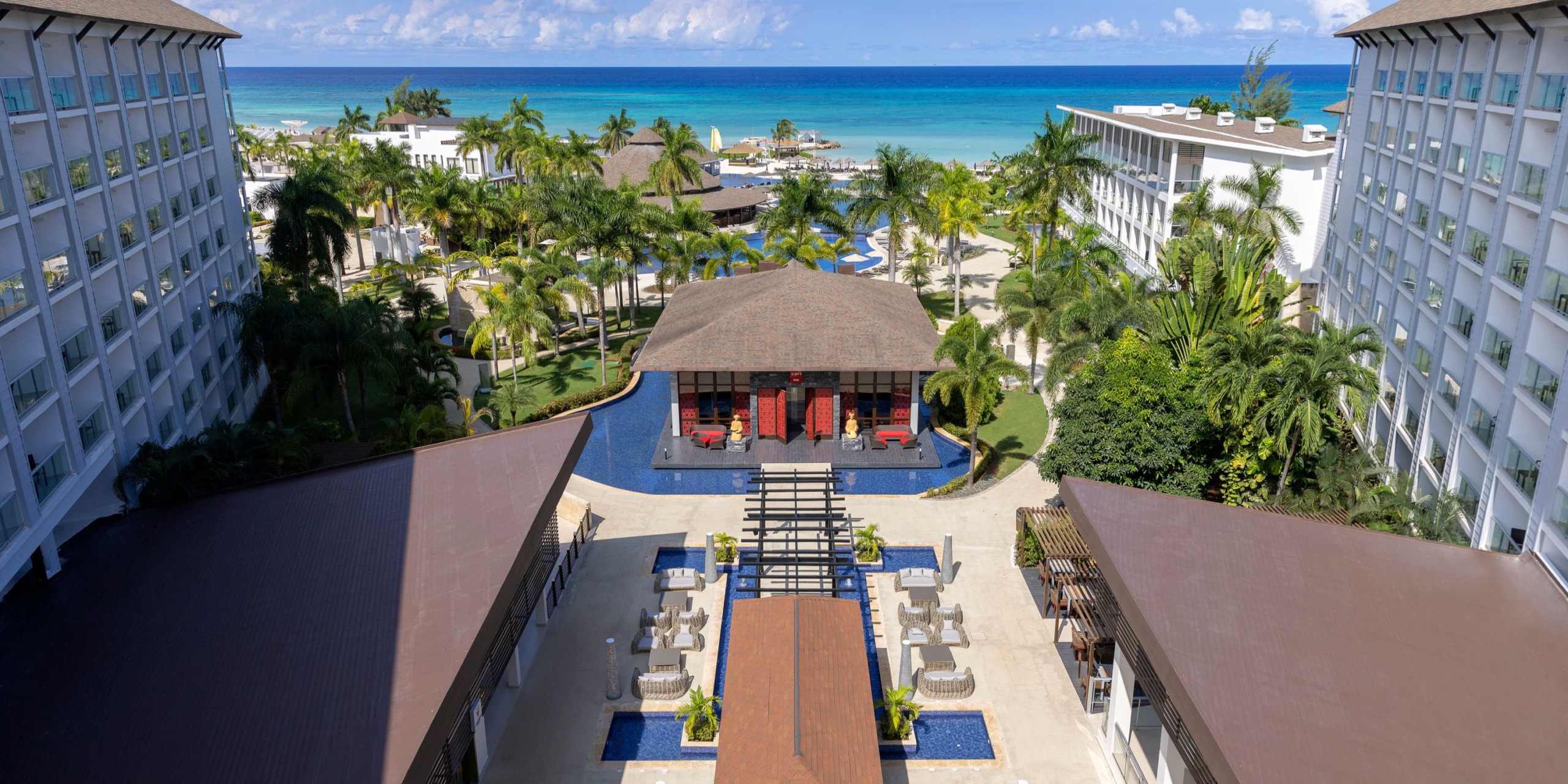 Aerial view of Hideaway at Royalton Blue Waters, showcasing two large hotel buildings, a central courtyard with a pool, surrounded by palm trees, and offering an ocean view in the background.
