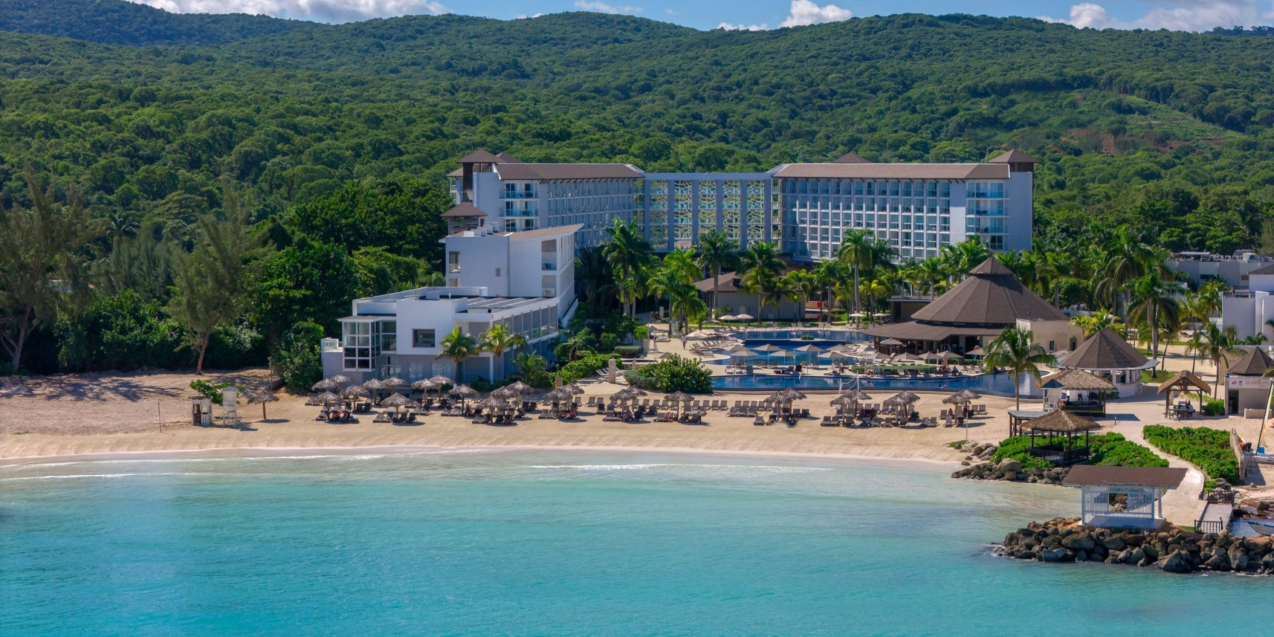 Hideaway at Royalton Blue Waters is a beachfront resort featuring multiple buildings, a pool, and sun loungers on the sand, with lush green hills as a stunning backdrop.