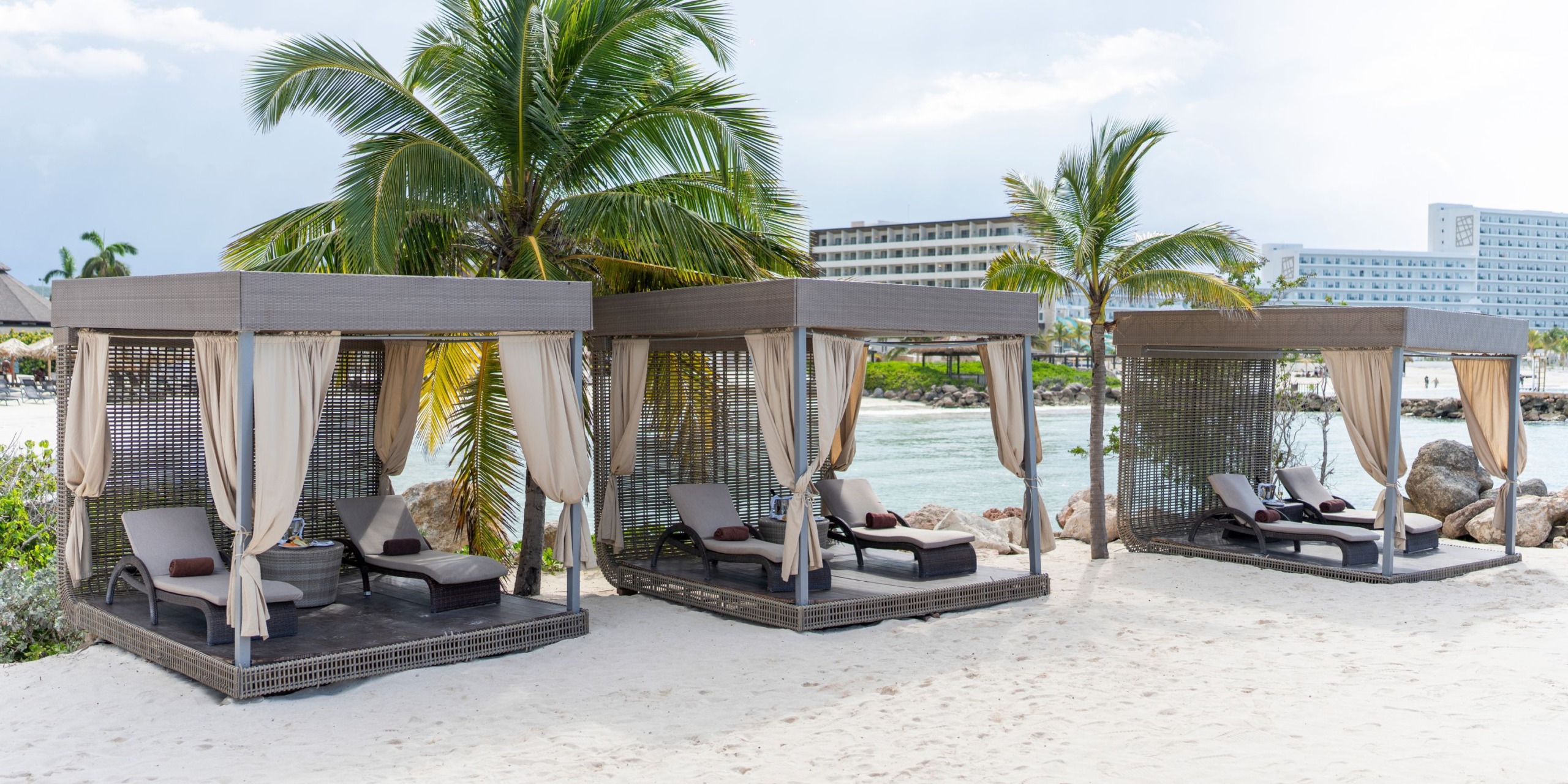 Three cabanas with lounge chairs on a sandy beach, surrounded by palm trees at Hideaway at Royalton Blue Waters. A resort building is visible in the background.