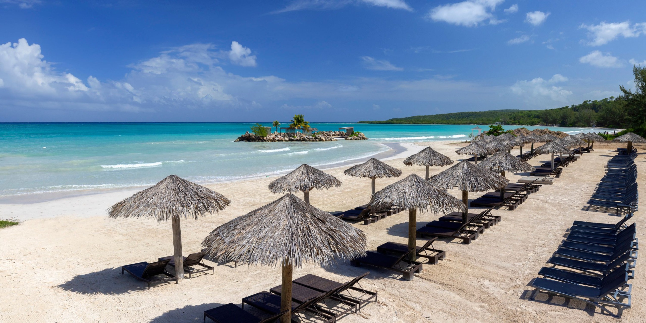 A tropical beach scene at Hideaway at Royalton Blue Waters features rows of thatched umbrellas and lounge chairs, overlooking turquoise water with a small island visible under a clear blue sky.