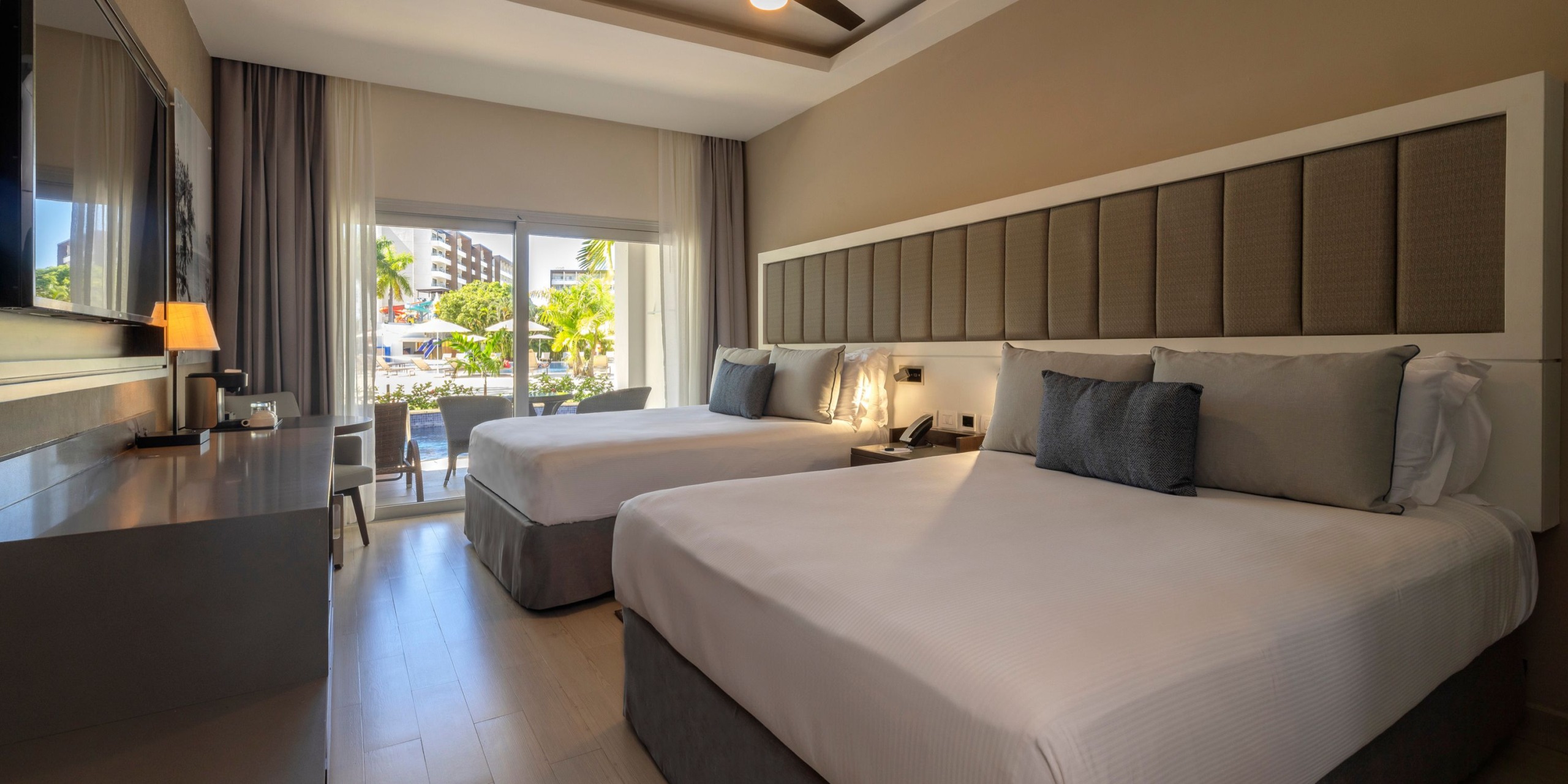 Room at Hideaway at Royalton Blue Waters featuring two neatly made queen beds, a large window overlooking trees and buildings, a desk with a chair, and a wall-mounted TV.