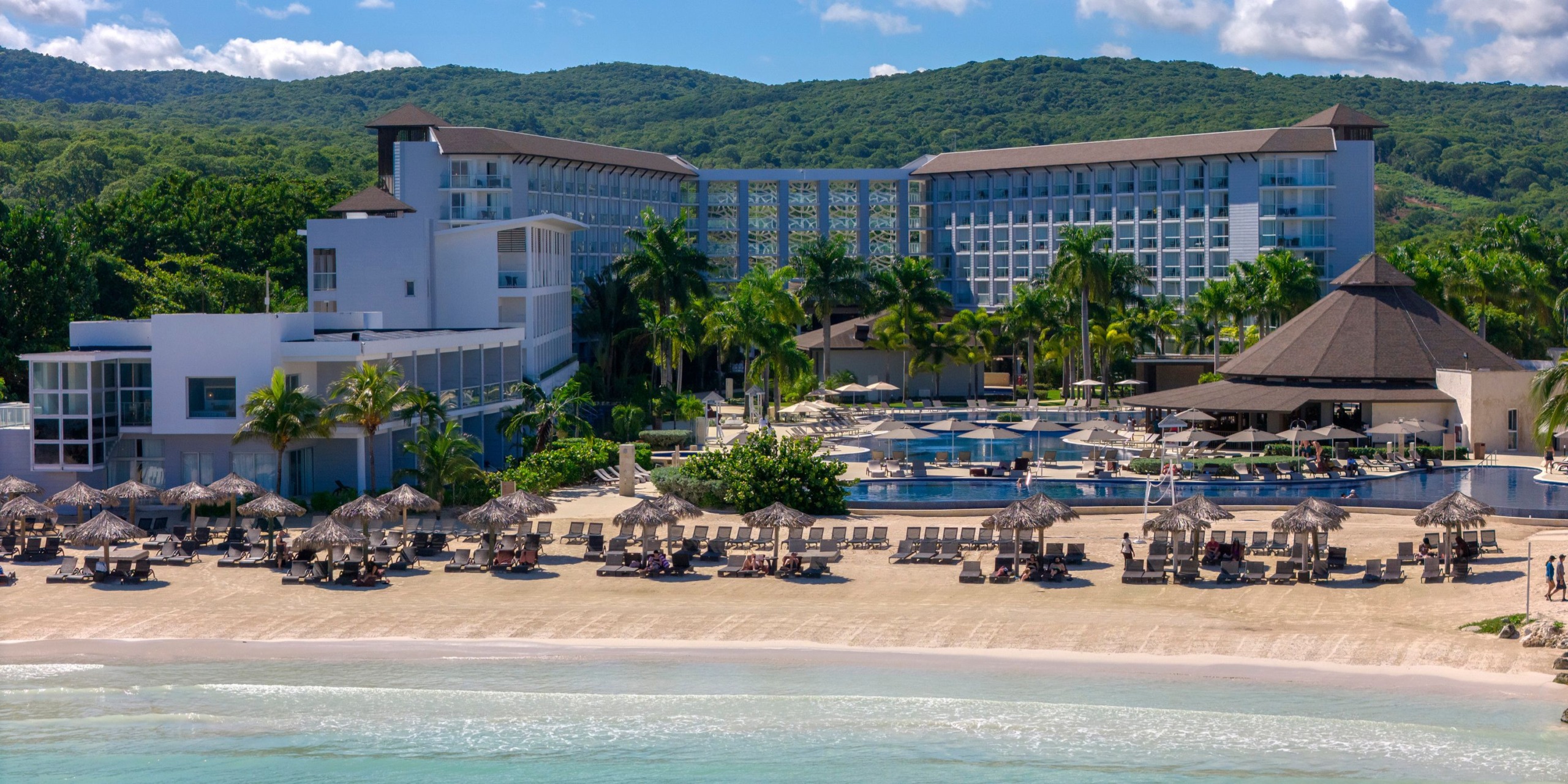 Hideaway at Royalton Blue Waters is a beachfront resort featuring two spacious hotel buildings, a pool area complete with umbrellas and sun loungers, all set amidst lush greenery with scenic mountains in the backdrop.