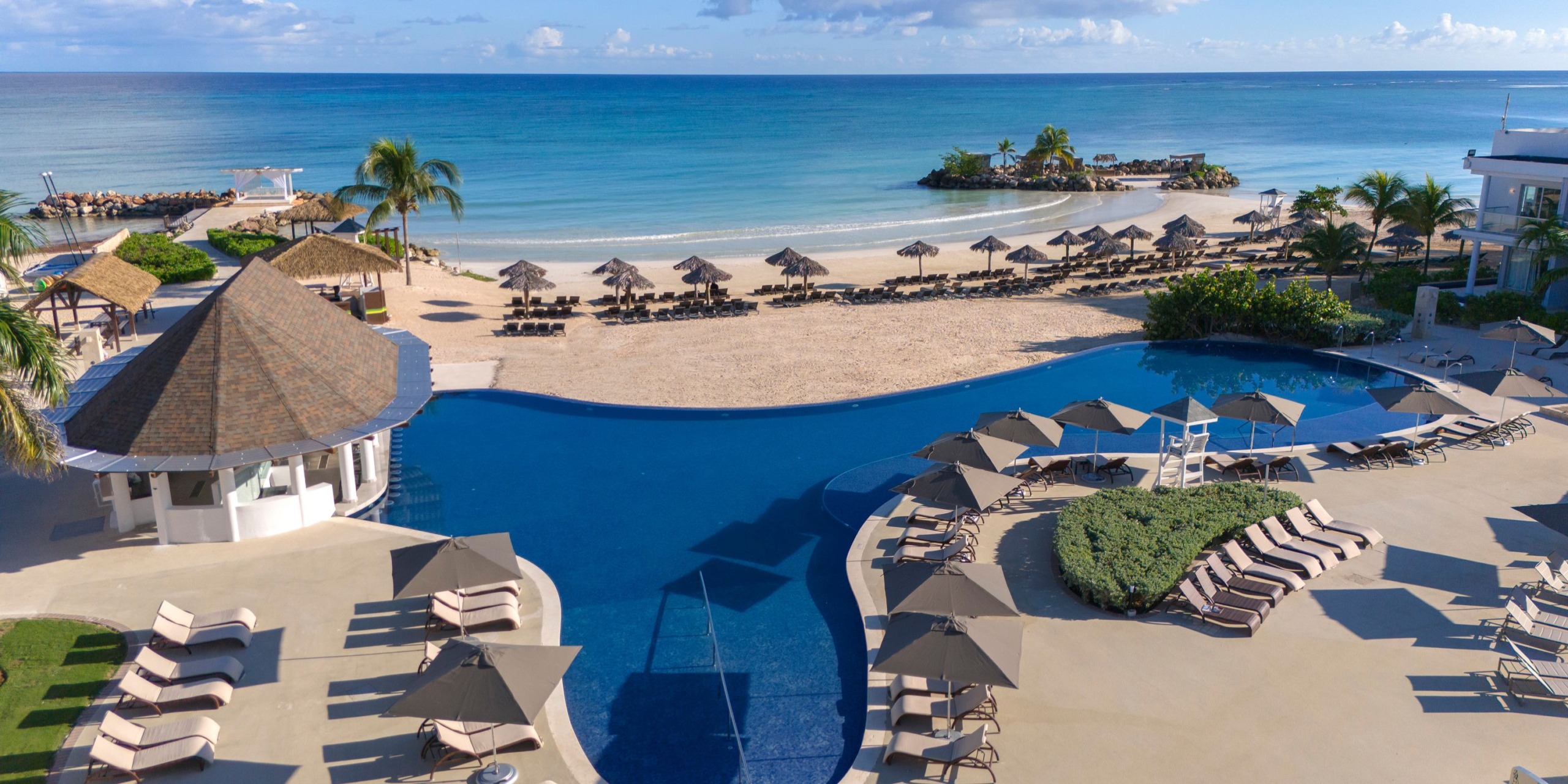 Aerial view of the Hideaway at Royalton Blue Waters resort, showcasing a large pool, sun loungers, umbrellas, and a sandy beach lined with more loungers and palm trees. The ocean appears calm beneath a clear sky.