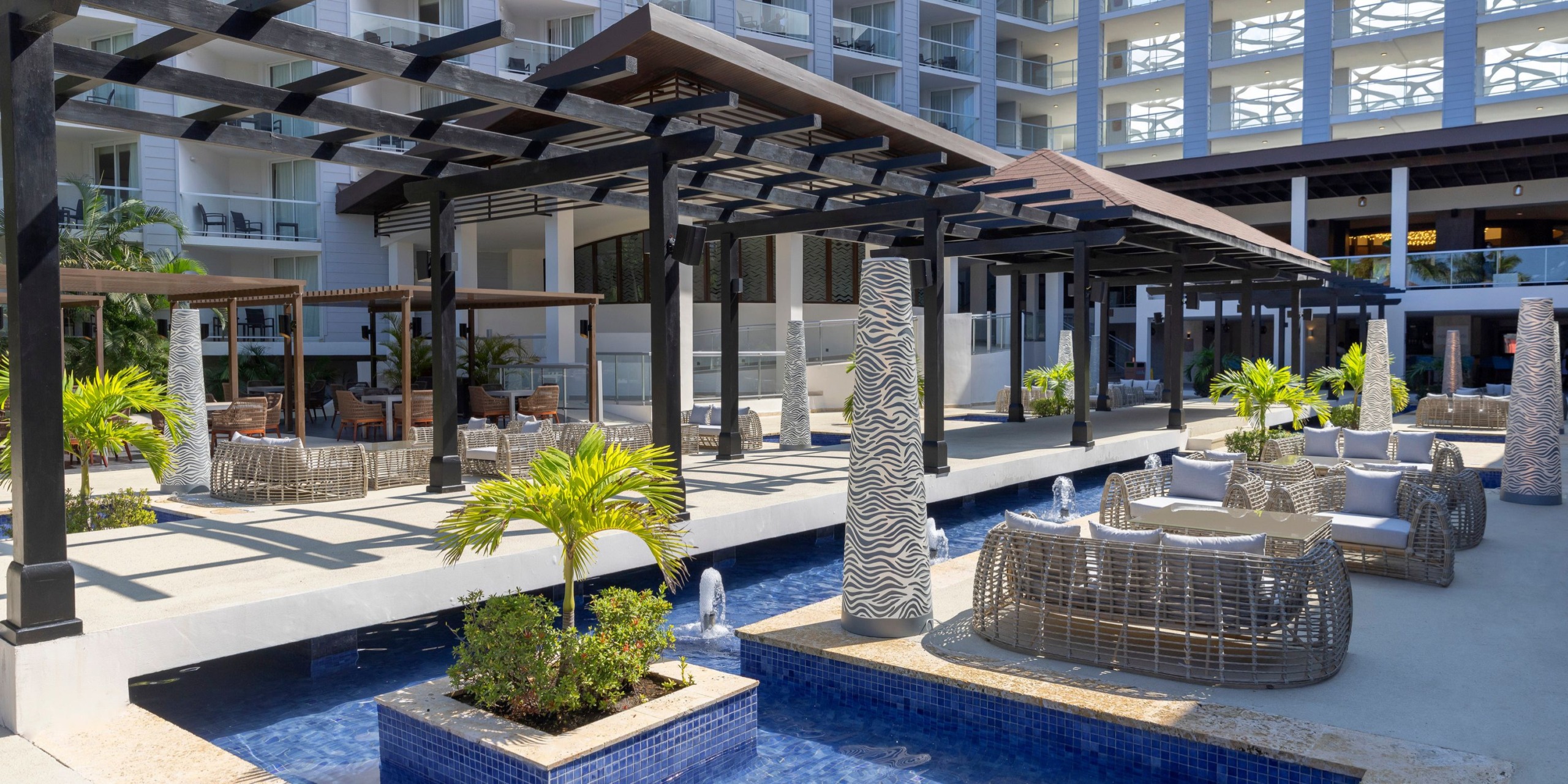 Outdoor seating area featuring wicker furniture and pergolas beside a shallow pool at the Hideaway at Royalton Blue Waters, a modern hotel.