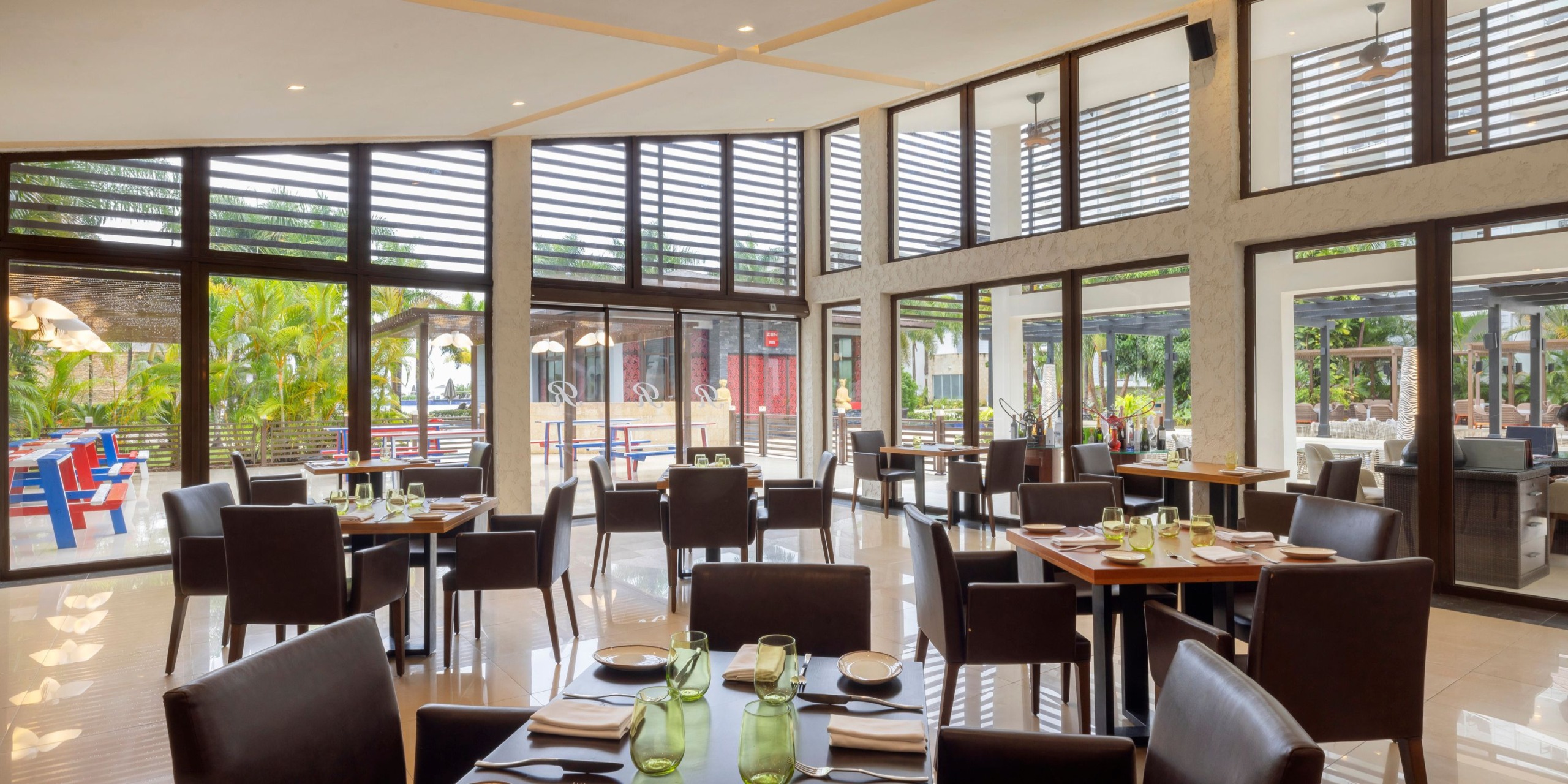 Introducing the Hideaway at Royalton Blue Waters: a spacious dining venue with contemporary decor, complete with wooden tables and chairs. Large windows flood the space with natural light, offering views of lush greenery outside.