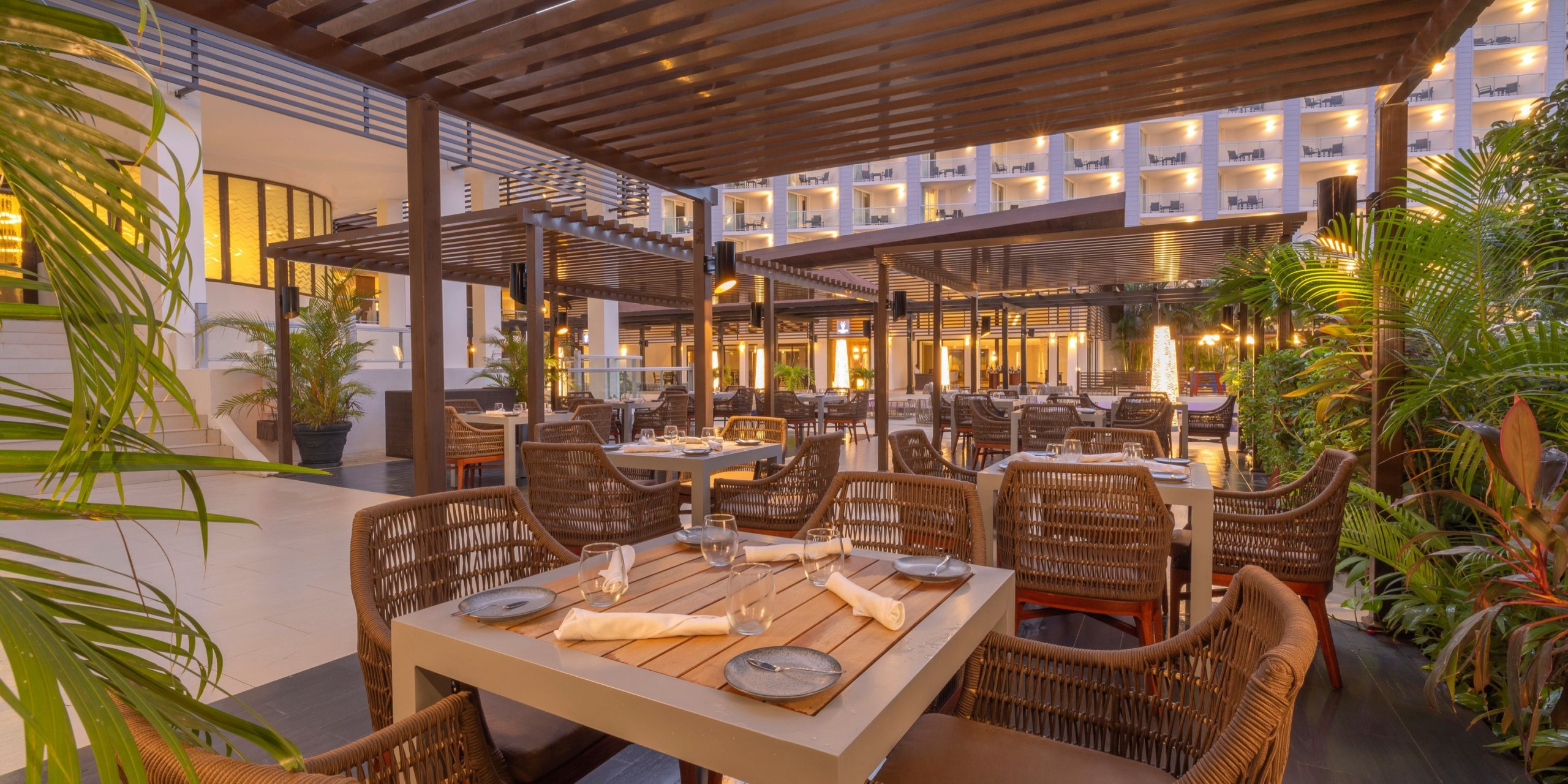 The outdoor dining area at Hideaway at Royalton Blue Waters features wicker chairs and wooden tables with stylish place settings under elegant pergolas, while a beautifully lit hotel building serves as a stunning backdrop.