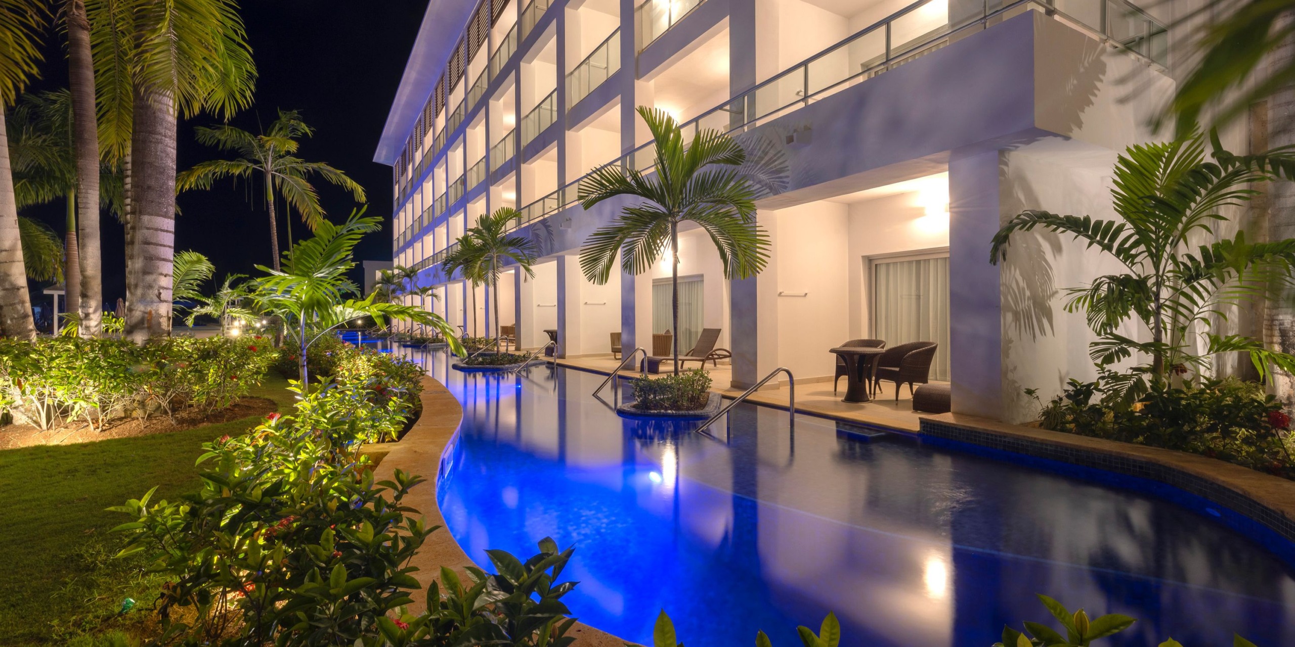 Night view of the Hideaway at Royalton Blue Waters, featuring illuminated balconies and a pool surrounded by lush tropical plants in the foreground.