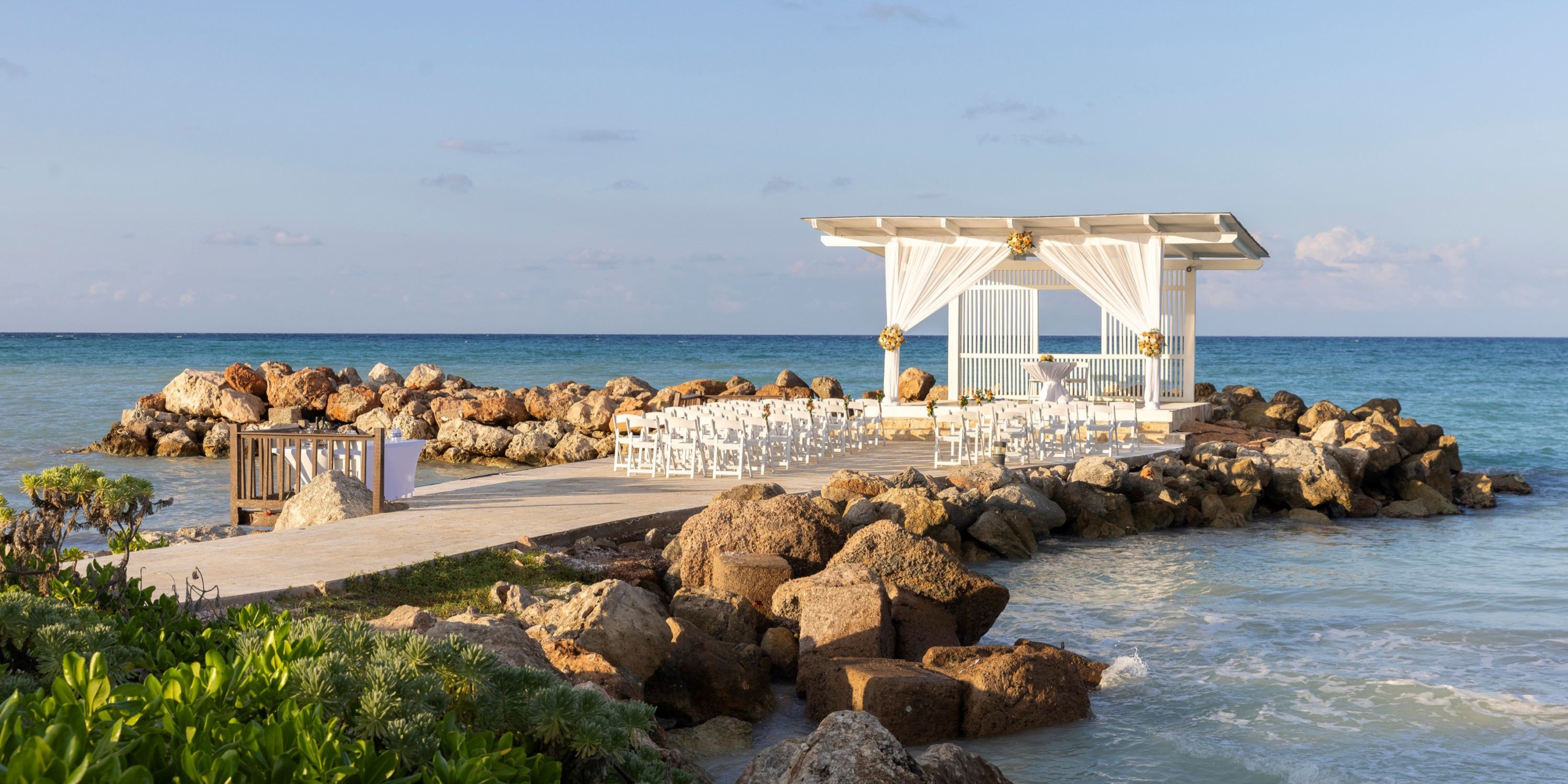 Hideaway at Royalton Blue Waters offers a beachfront wedding venue featuring a white pergola perched on rocks by the sea, with rows of chairs arranged for guests.