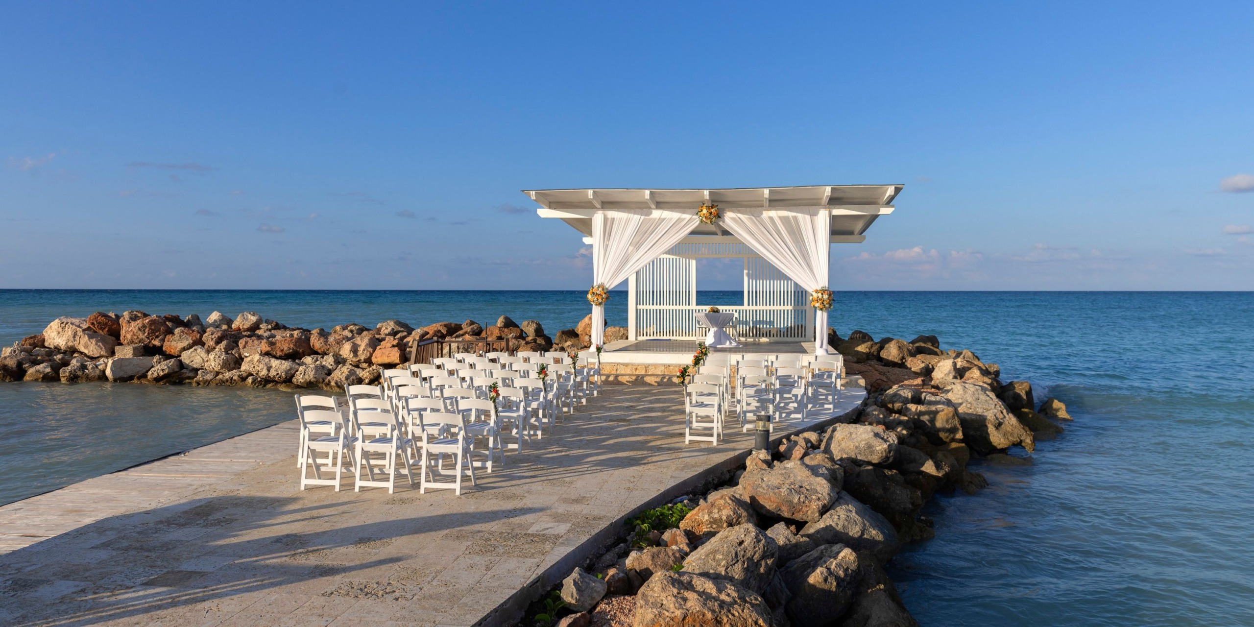 The Hideaway at Royalton Blue Waters offers an outdoor wedding venue set up on a stone pier, overlooking the ocean with rows of white chairs and a beautifully decorated pergola under clear blue skies.