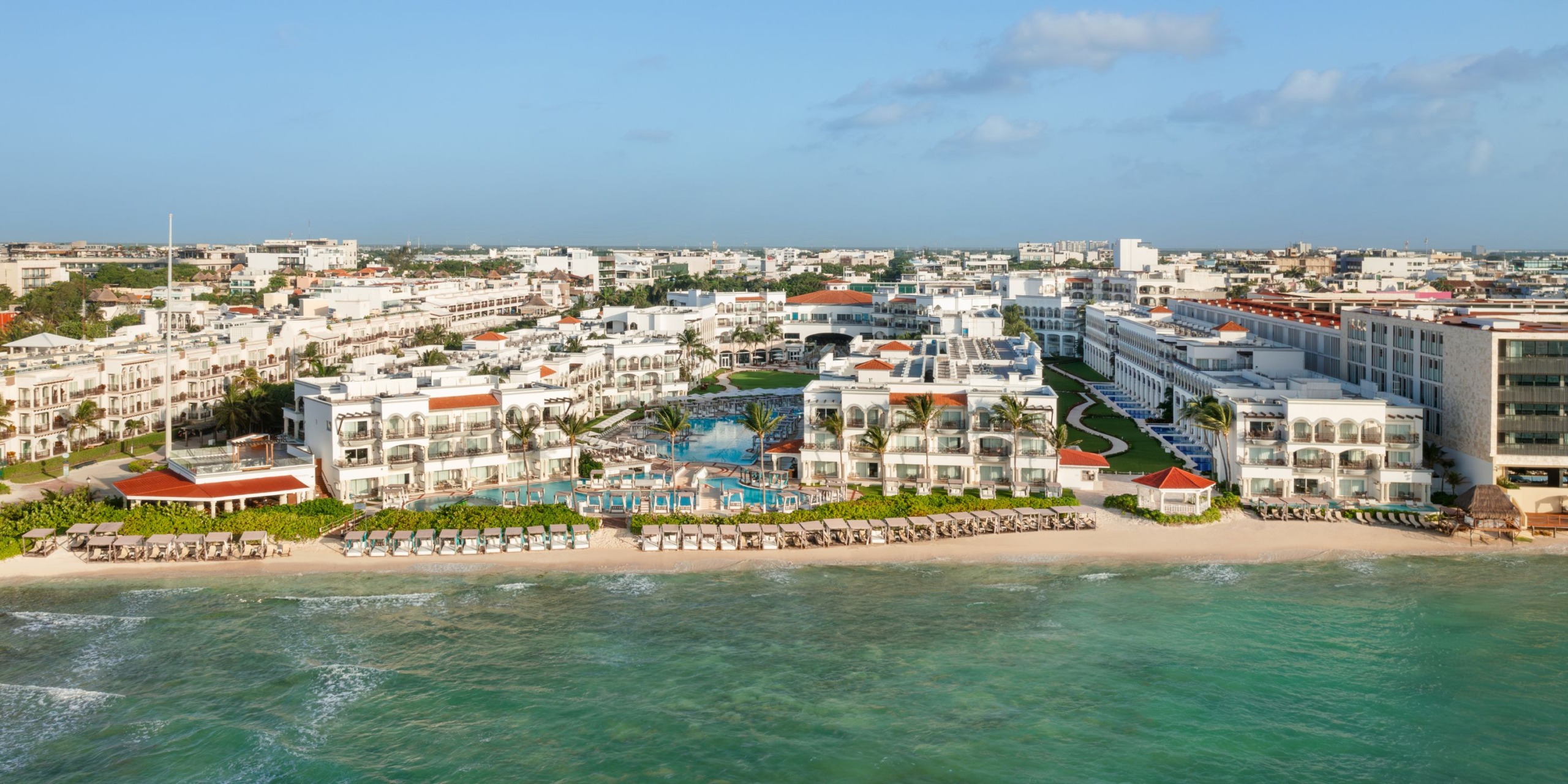 Aerial view of Hilton Playa del Carmen, a beachfront resort featuring multiple buildings, a central pool area, sun loungers, and umbrellas along the shore. The ocean is visible in the foreground.