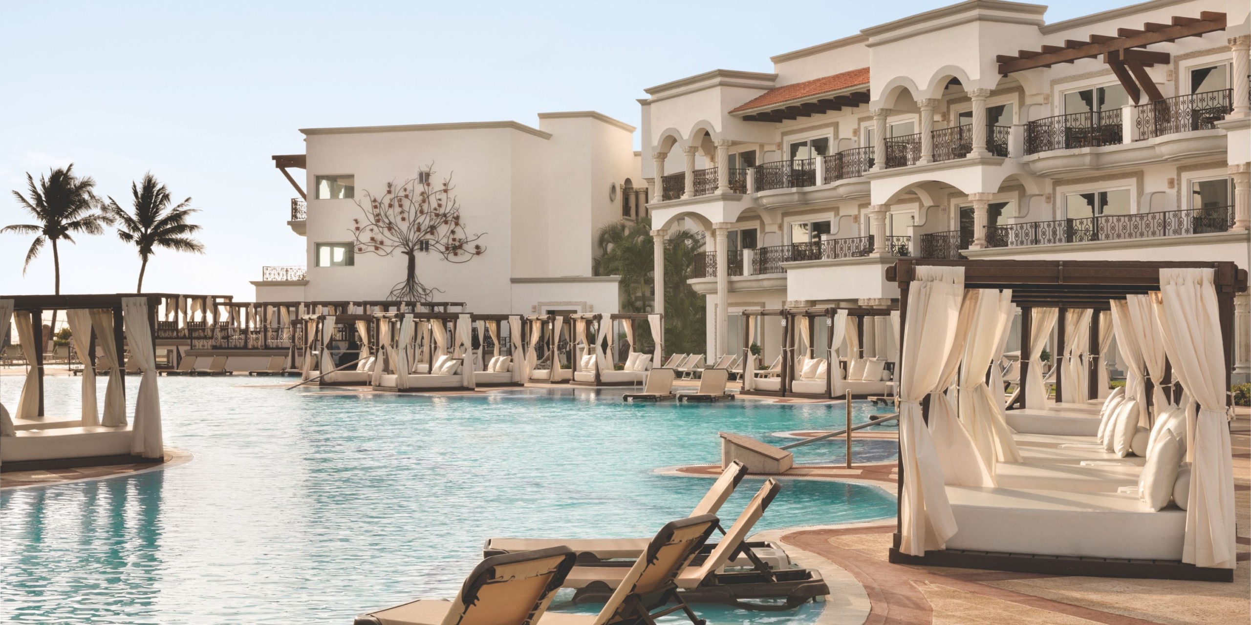 The Hilton Playa del Carmen features a resort pool area complete with lounge chairs and cabanas, all set under a clear sky and encircled by elegant multi-story buildings in a tropical setting.