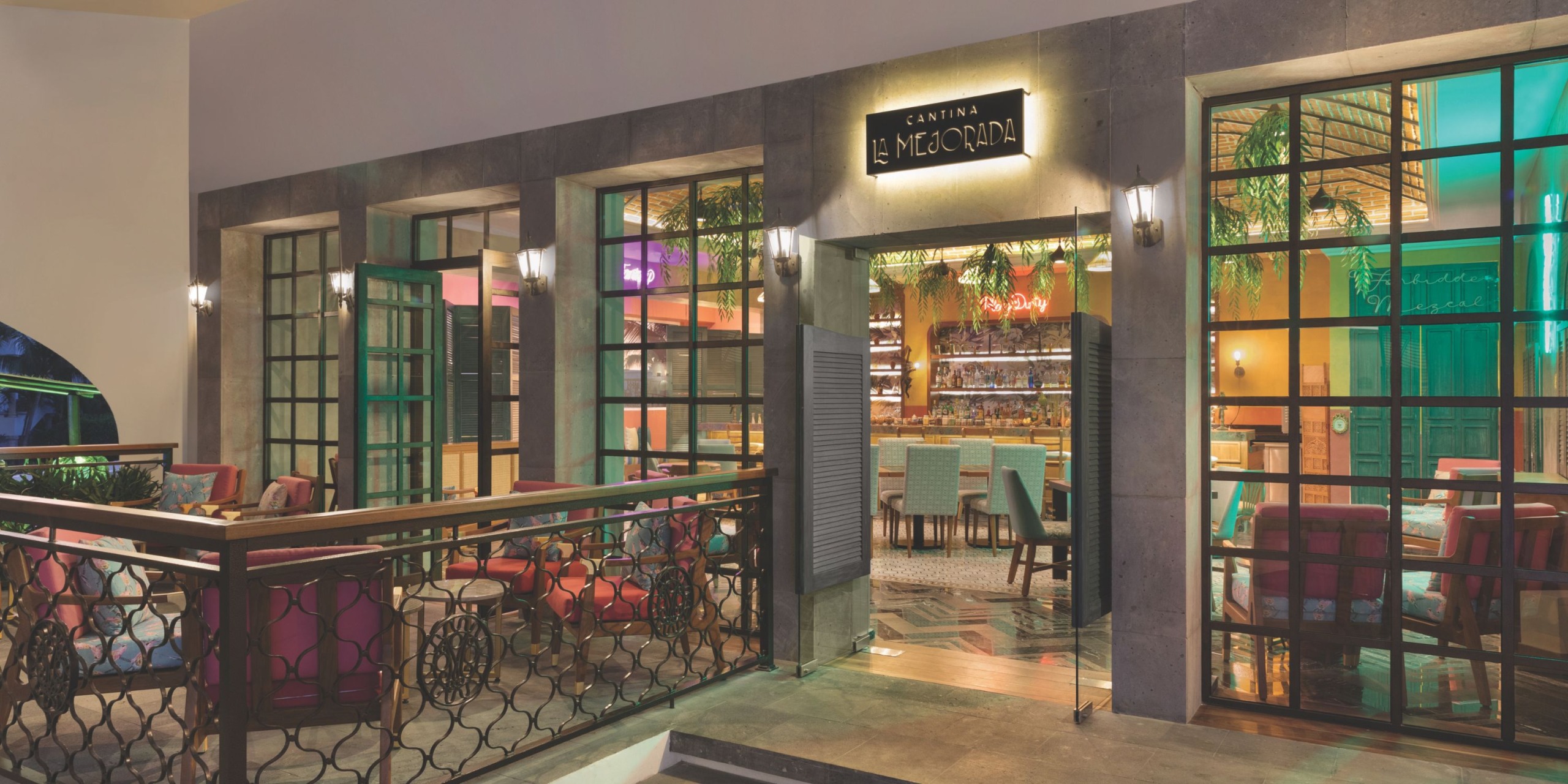 Outdoor view of a cantina with large windows showing colorful interior seating and a bar area, featuring a sign "Hilton Playa del Carmen" above the entrance.